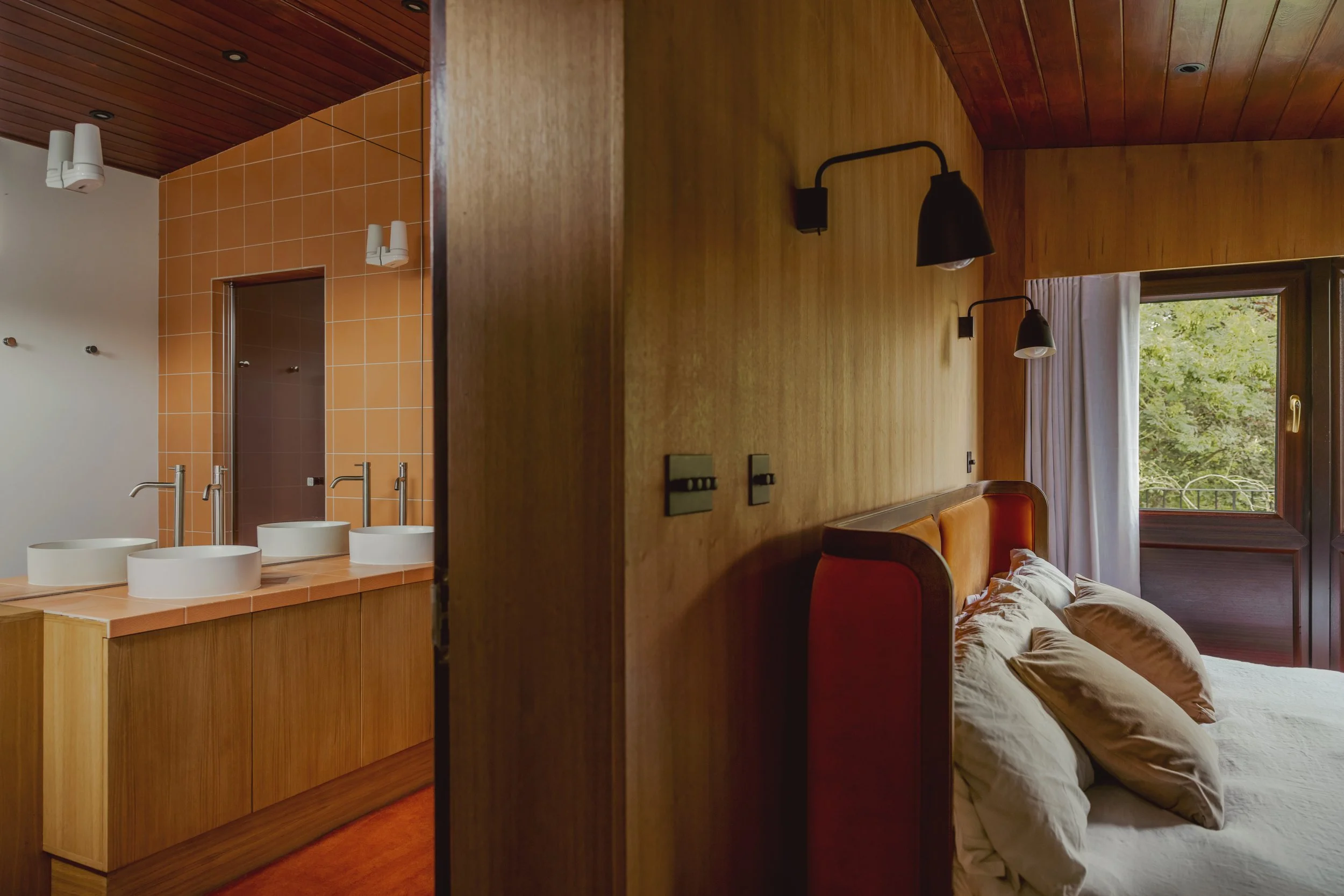 A bedroom with wooden walls, a bed with beige pillows, and a window overlooking green trees. Adjacent to the bedroom is a bathroom with three vessel sinks, orange tiles, and wood cabinetry.