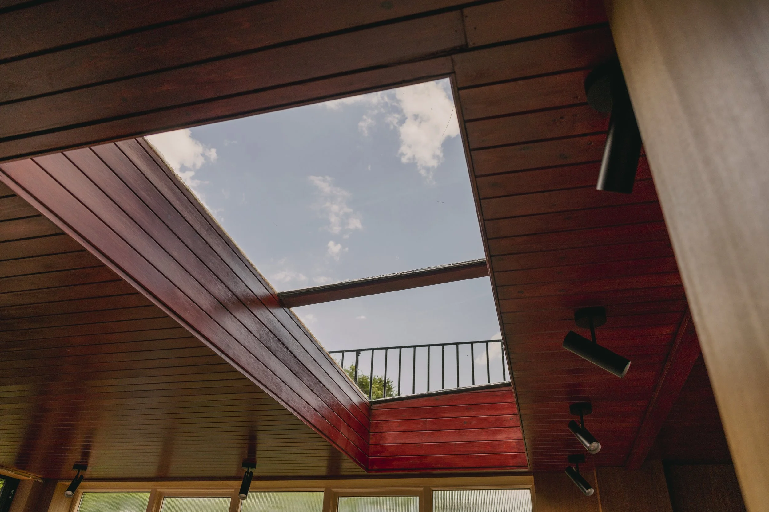 Open skylight in a wooden ceiling showing blue sky and clouds, with black track lighting.