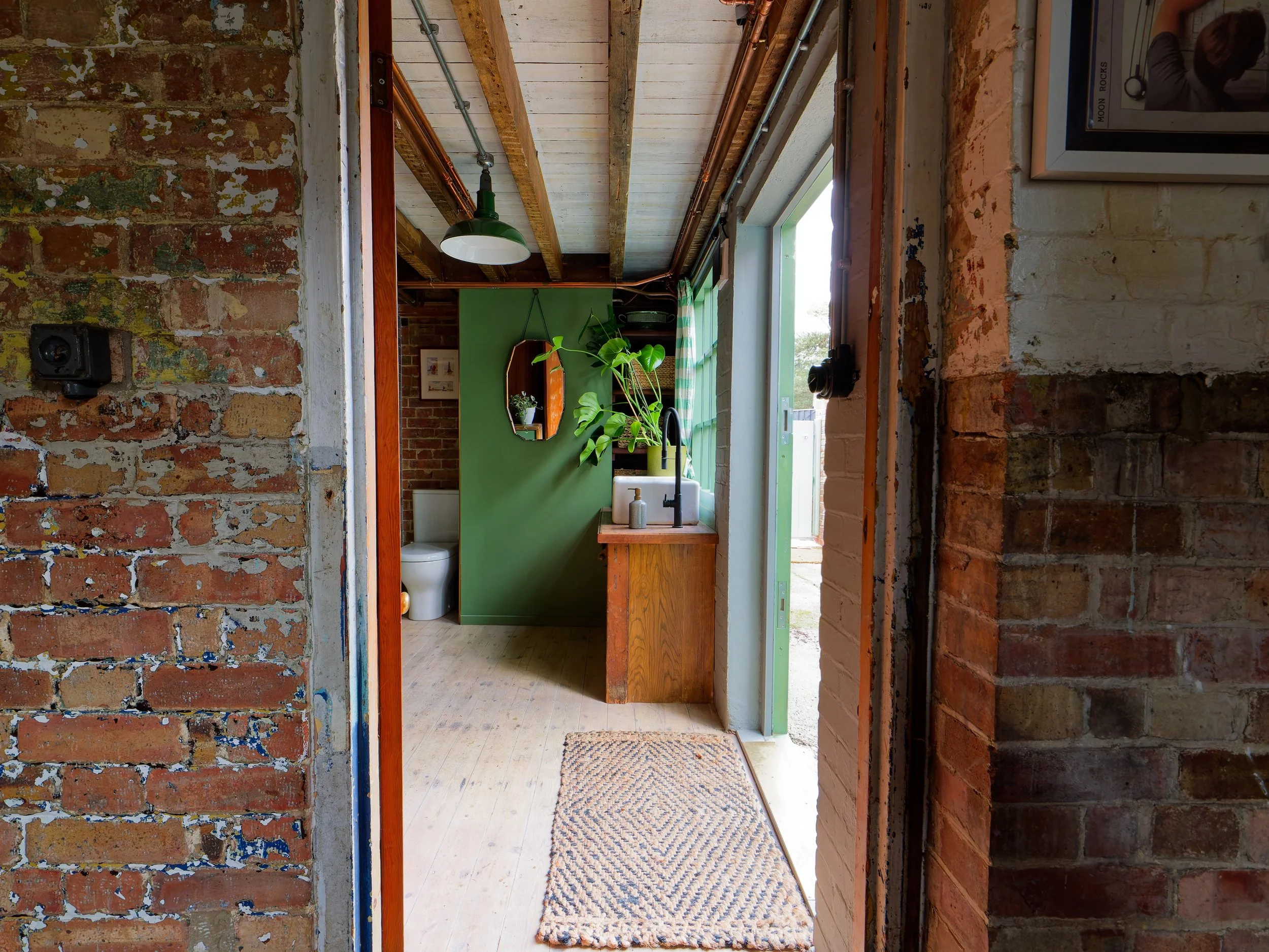 View through an entryway into a rustic kitchen with exposed brick walls, wooden ceiling, and green accent wall. A small wooden sink cabinet with a black faucet, potted plant, and soap dispensers near a window. A separate toilet room with a white toil