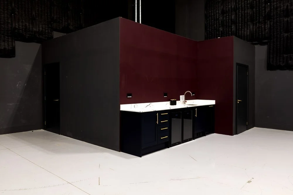 A small kitchenette with dark cabinets, a white marble countertop, and a sink, set against a dark red and black wall, in a large open space.