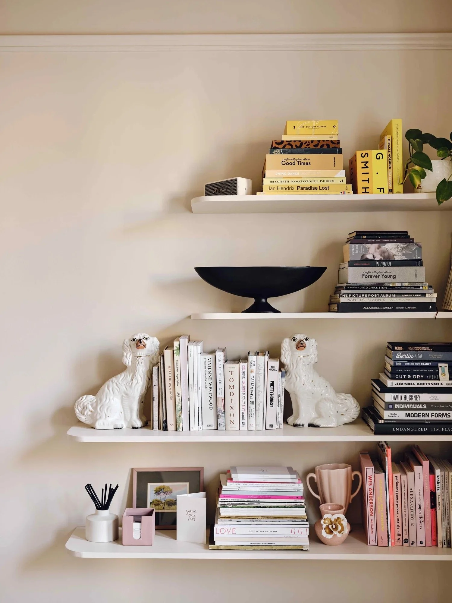 Three white floating shelves filled with books, decorative items, and plants against a beige wall.
