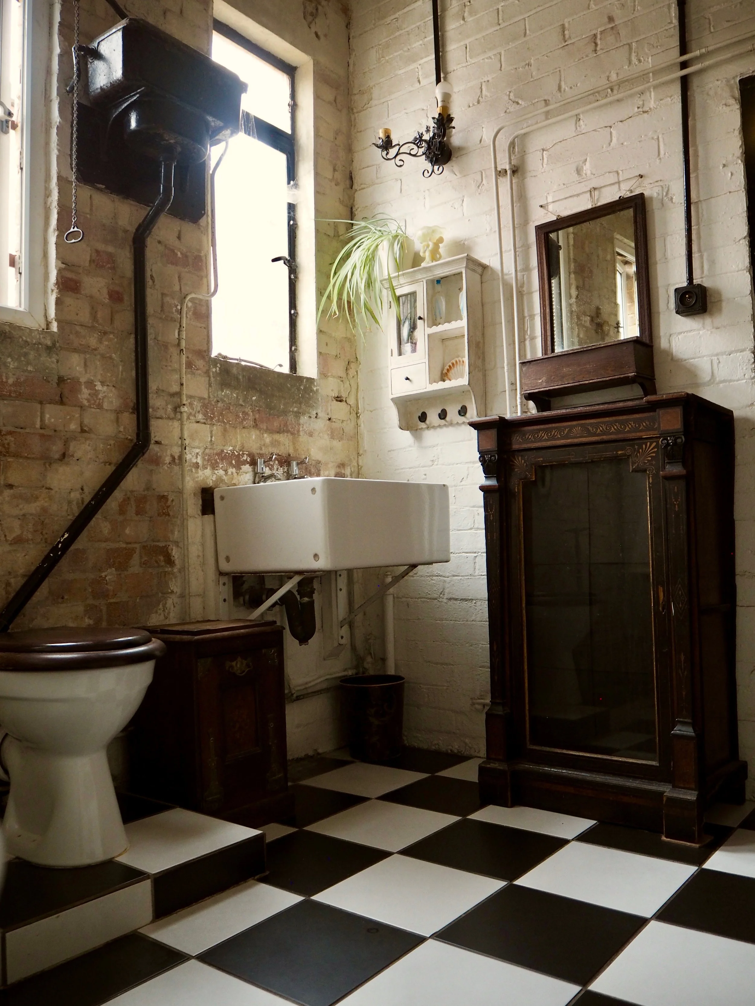 A bathroom featuring a checkered black and white tile floor, a toilet, a vintage wooden cabinet, a white wall-mounted sink, a small white wall cabinet with shelf and knobs, a window with natural light, a black wall-mounted telephone, and various deco