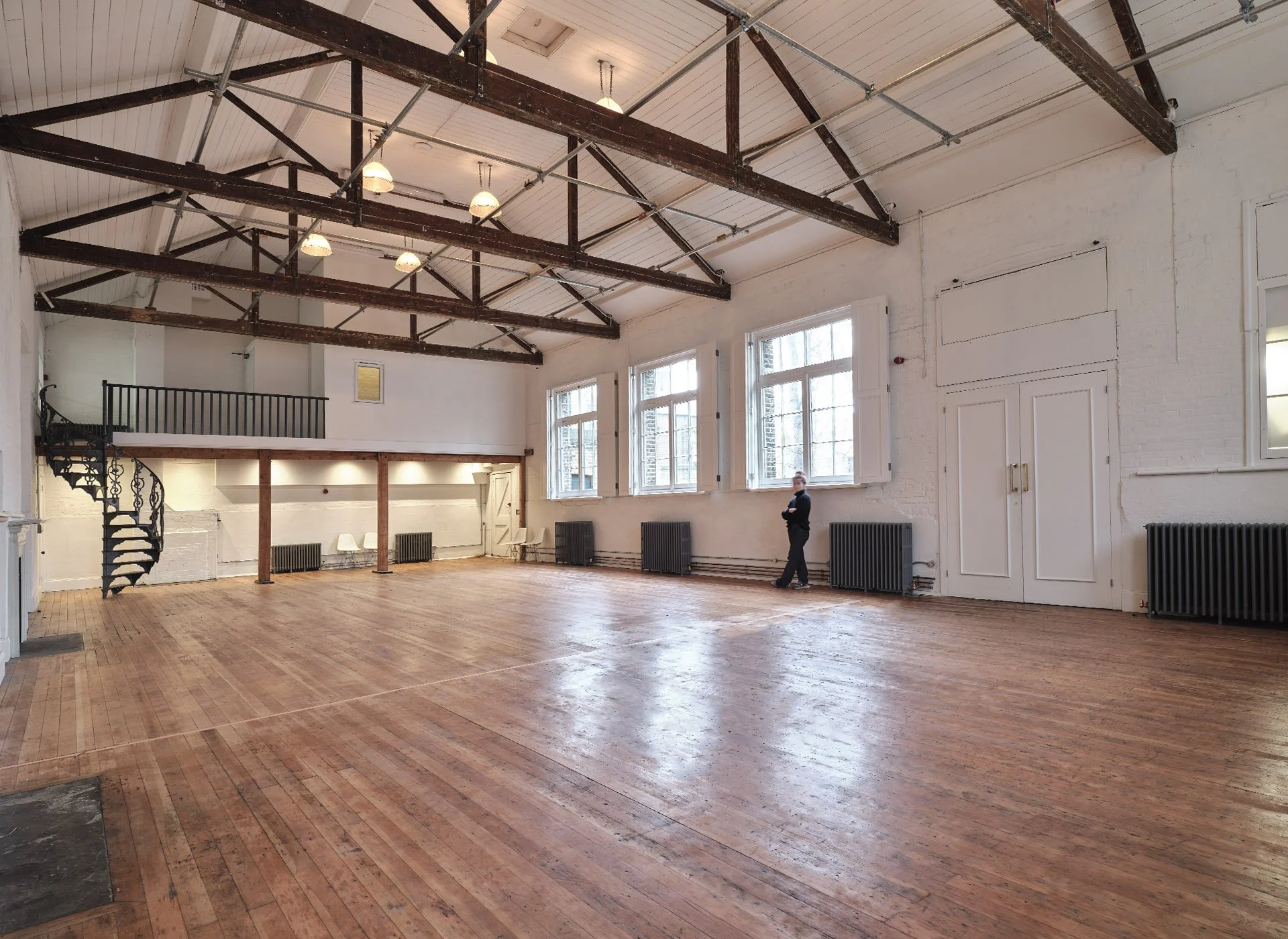 Spacious, well-lit room with wooden floors, white walls, high ceiling with exposed beams, large windows, black radiators, and a person standing near the windows.