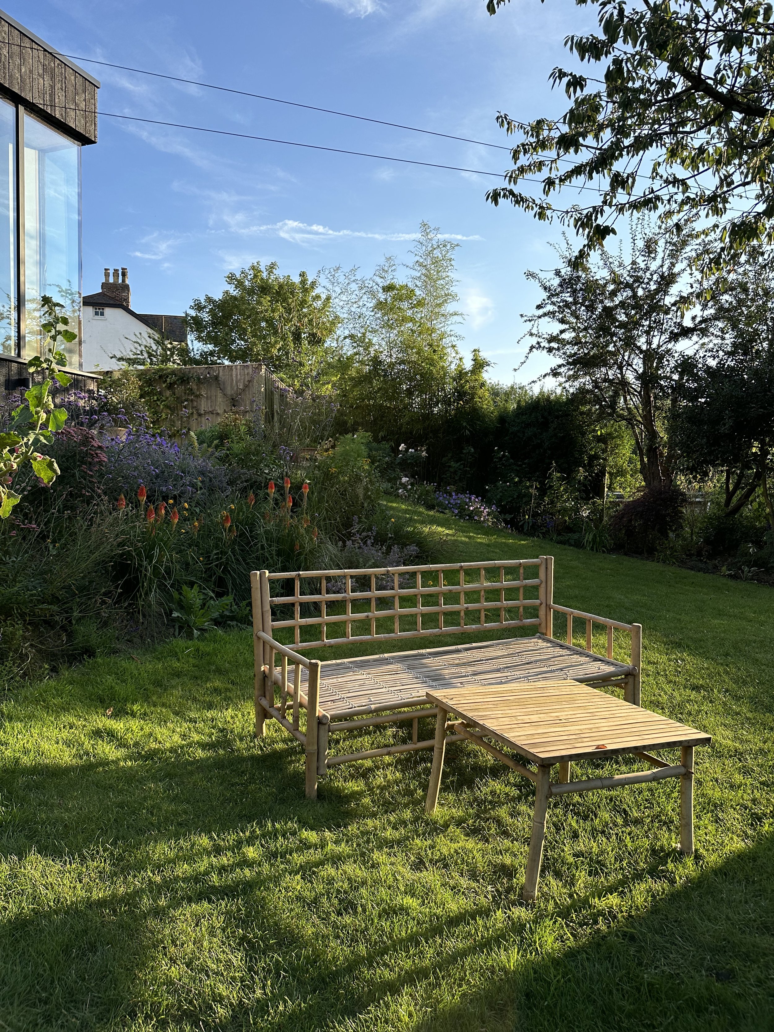 A wooden garden bench and small table on a grassy lawn in a lush garden with colorful flowers and trees, under a blue sky with some clouds.
