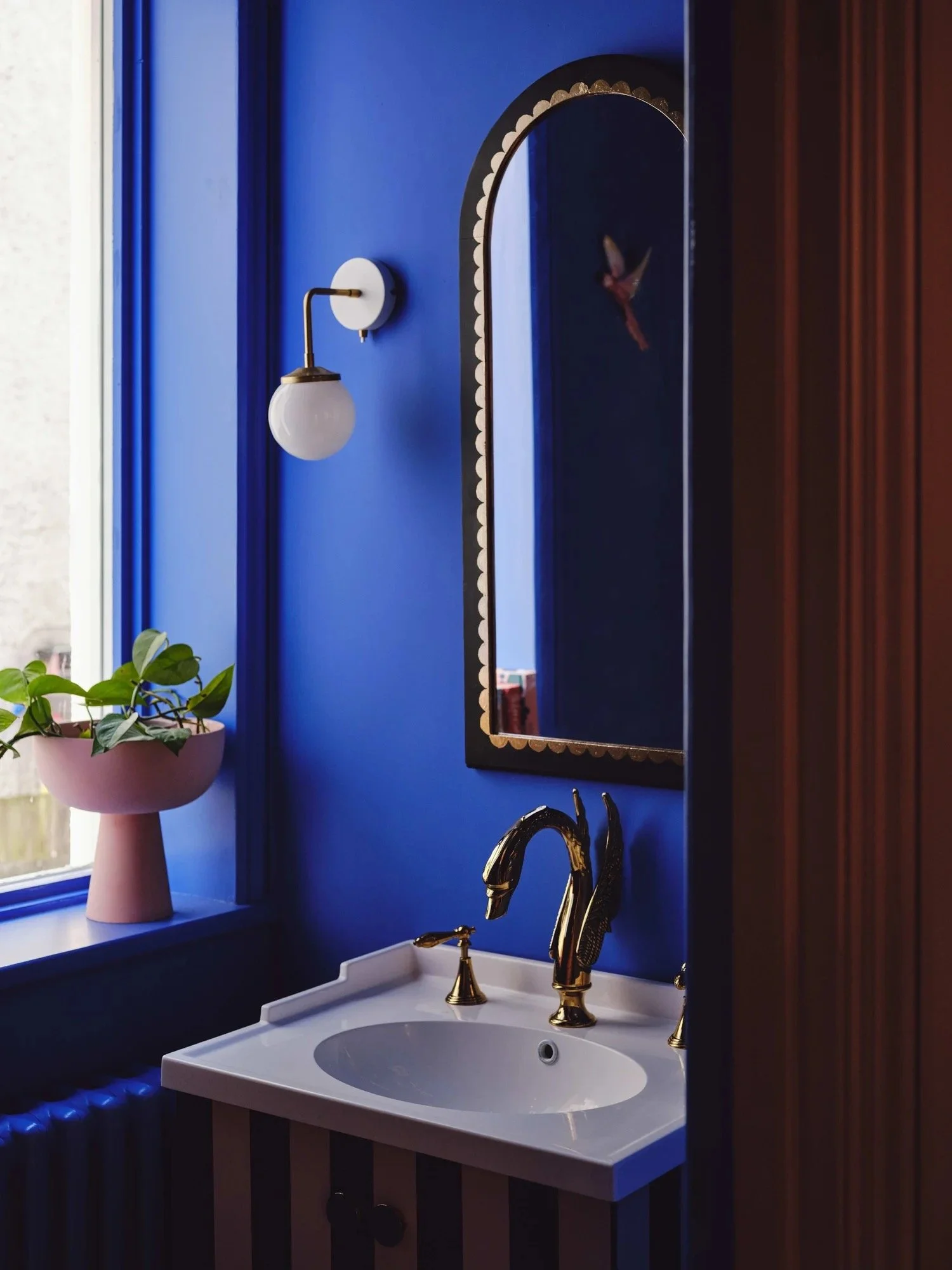 A bathroom with a deep blue wall, a white sink with gold fixtures, a black framed mirror with decorative gold trim, a wall-mounted light fixture with a white globe, a pink planter with green plants on the window sill, and a reflection of the hummingb