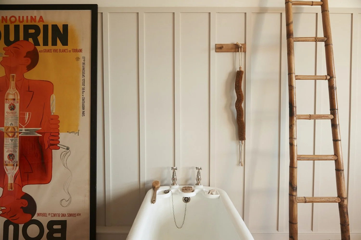Vintage bathtub with silver taps, a brush, and soaps on the edge, against a white paneled wall with a wooden ladder and a hanging scrub brush beside a framed vintage advertisement poster.