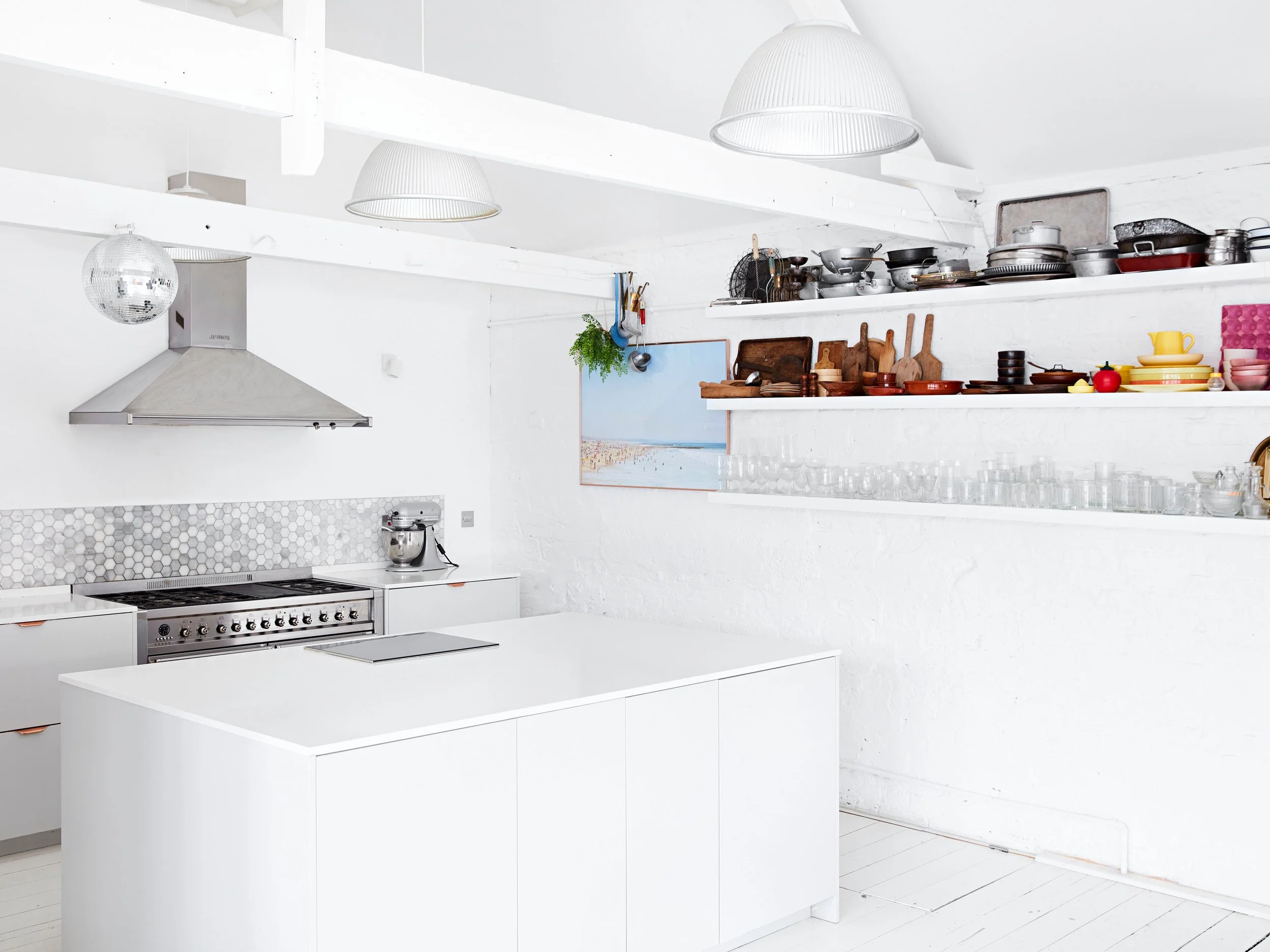 A modern, all-white kitchen with a central island, stainless steel appliances, open shelves with various kitchenware, and a artwork of a beach scene on the wall.
