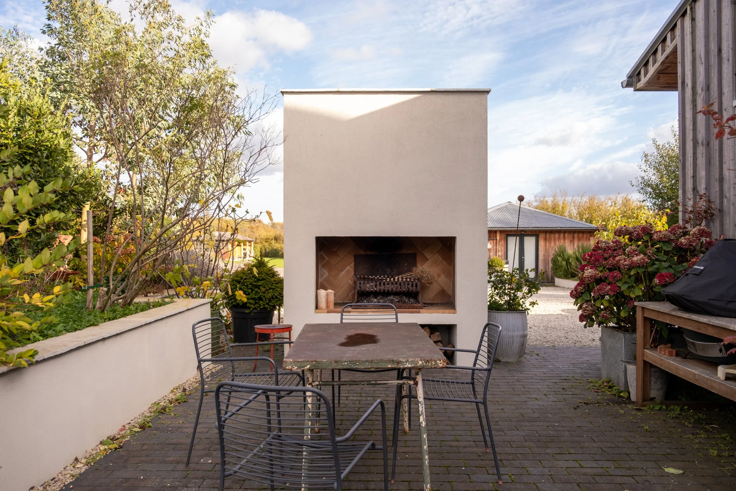 An outdoor patio area with a white fireplace, a table with chairs, surrounded by plants and trees, and a gravel pathway, with a wooden building in the background.