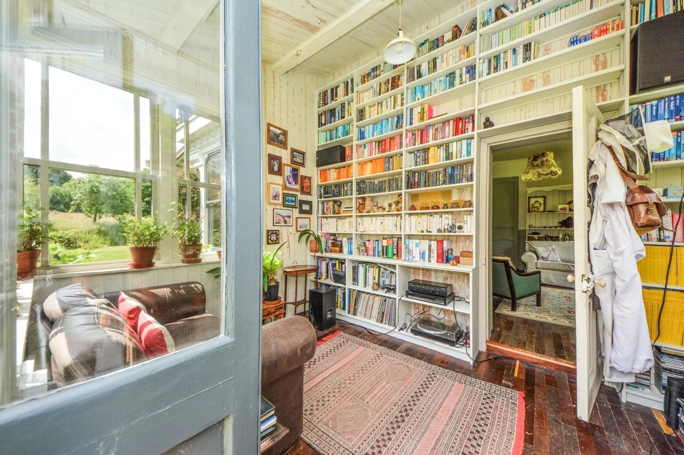 A cozy room with a large white bookshelf filled with books, a small sitting area with a brown sofa and red pillows, a window with potted plants outside, and a doorway leading to another room with more furniture.
