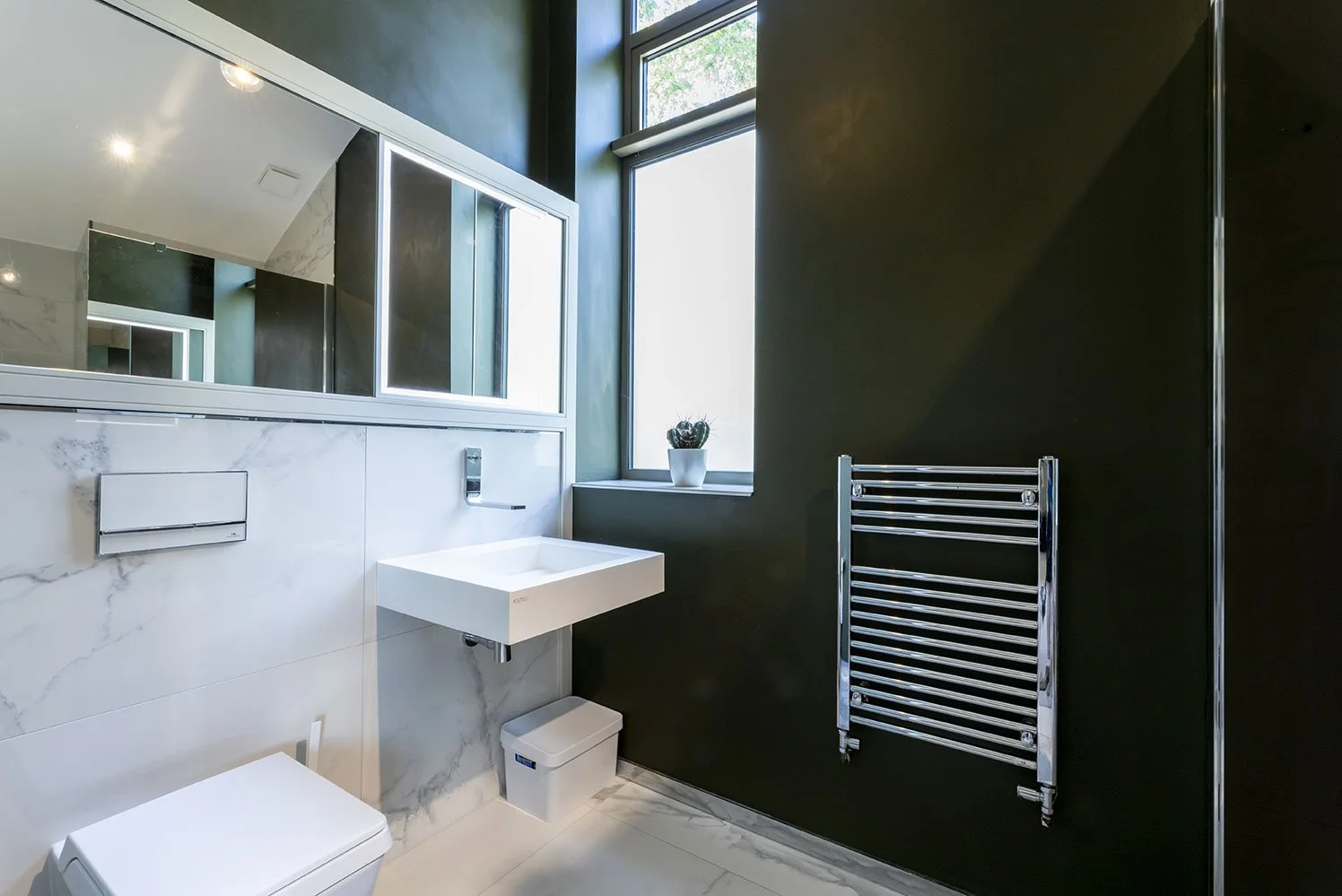 Modern bathroom with black walls and white marble accents, including a wall-mounted sink, a large mirror, a window with a potted plant, and a chrome towel warmer.