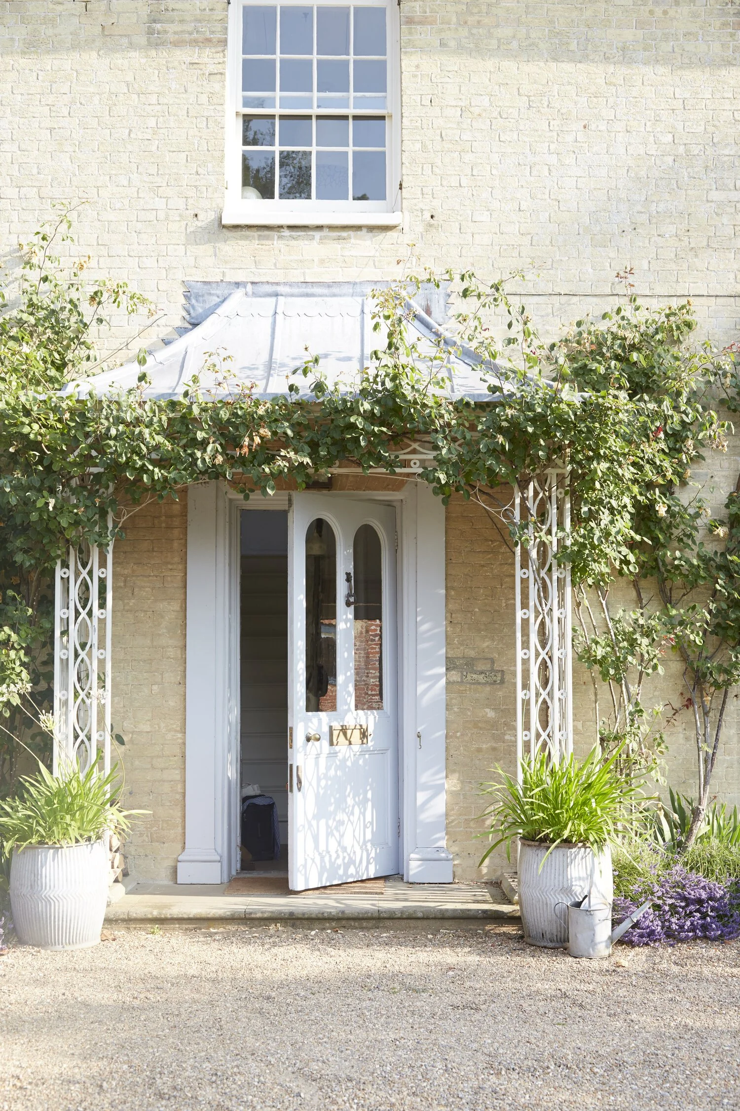 White front door with an arched window, open to a staircase inside, surrounded by two large white flower pots with green plants. A vine with green leaves and pink flowers climbs on a white metal trellis on each side of the door. Above the door is a s