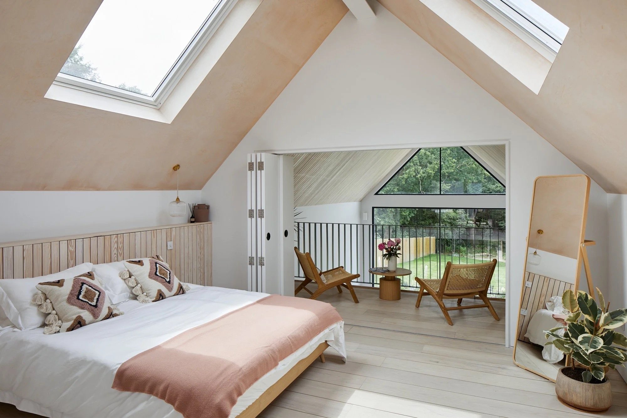 Bedroom with sloped ceiling, skylights, and a balcony with outdoor seating. The bed has a light wood headboard and decorative pillows, and a mirror stands beside a potted plant.