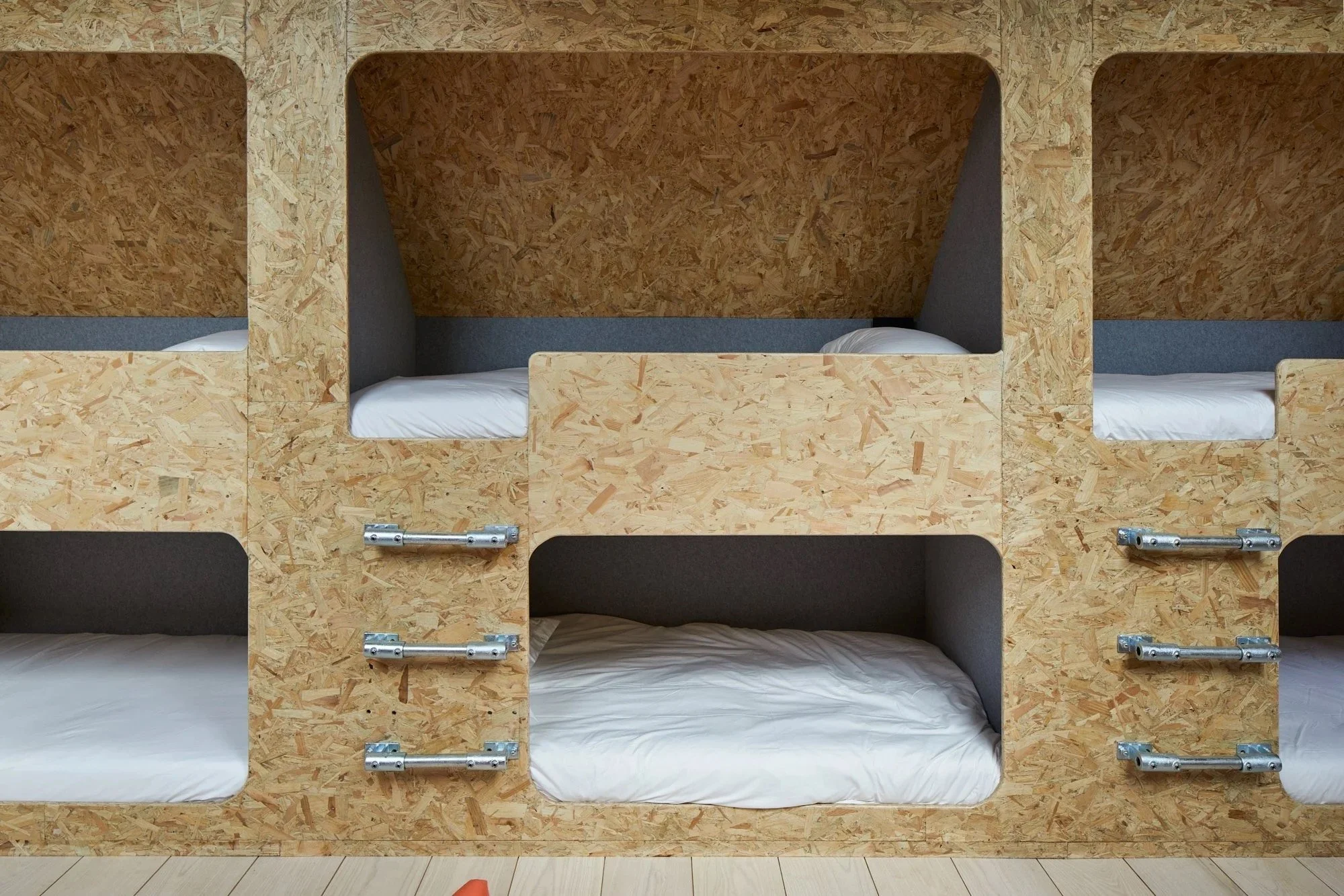 Multiple bunk beds made of plywood with white bedsheets and pillows, arranged in a modern, minimalist style.