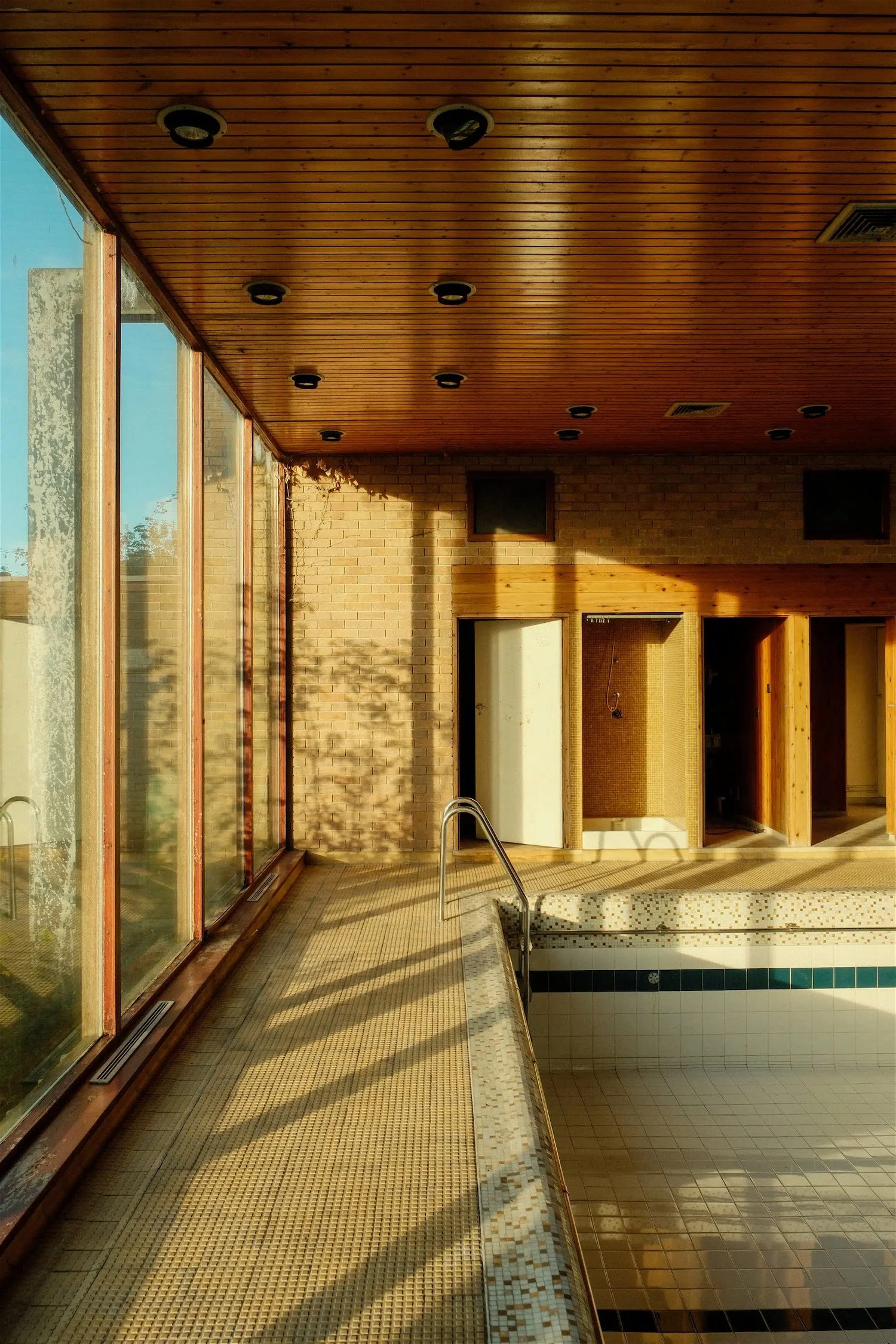 Empty indoor swimming pool area with sunlight streaming through large glass windows, brick walls, wooden ceiling, and tiled flooring.