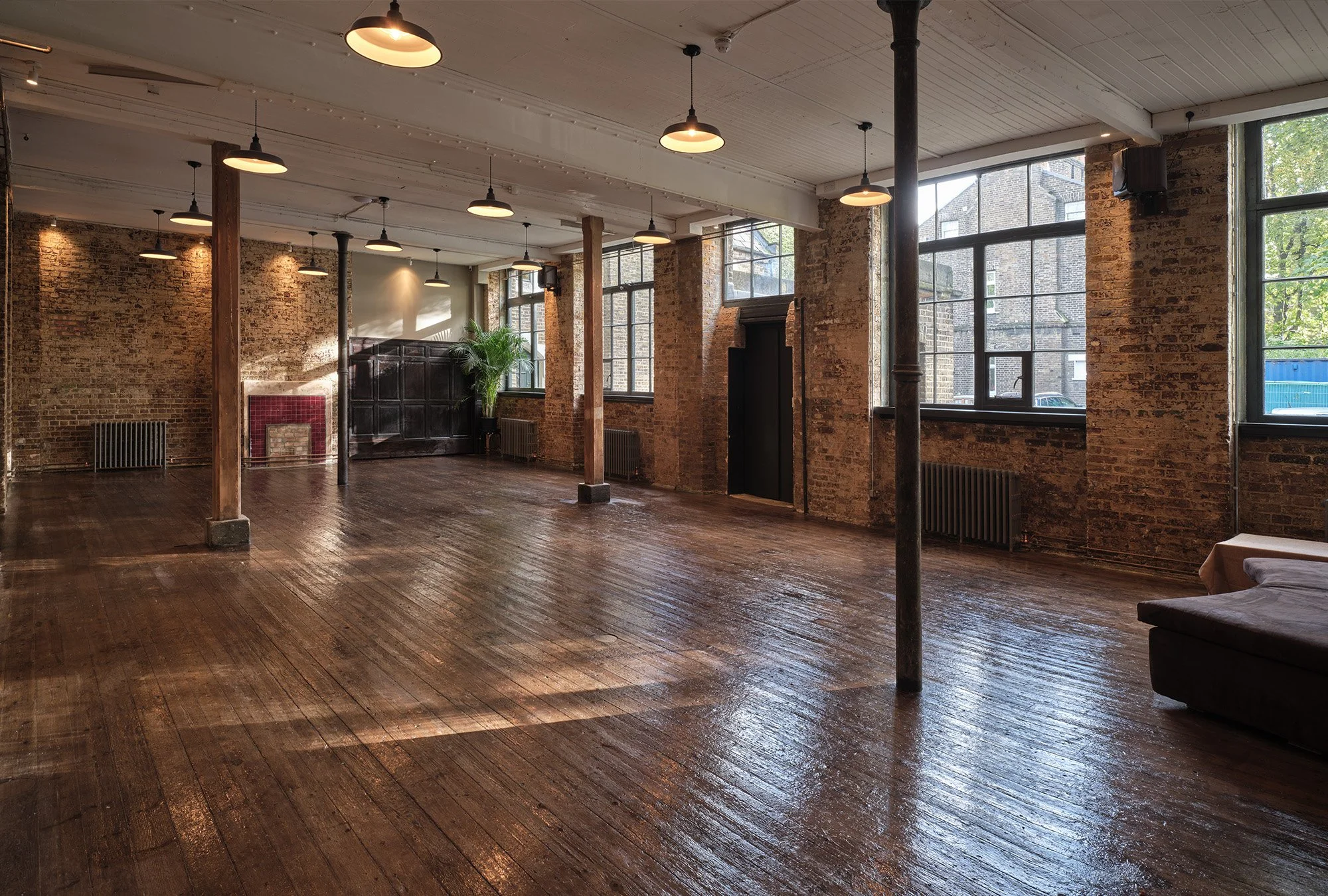 Empty industrial-style room with large windows, exposed brick walls, wooden floors, hanging pendant lights, and a few pieces of furniture.