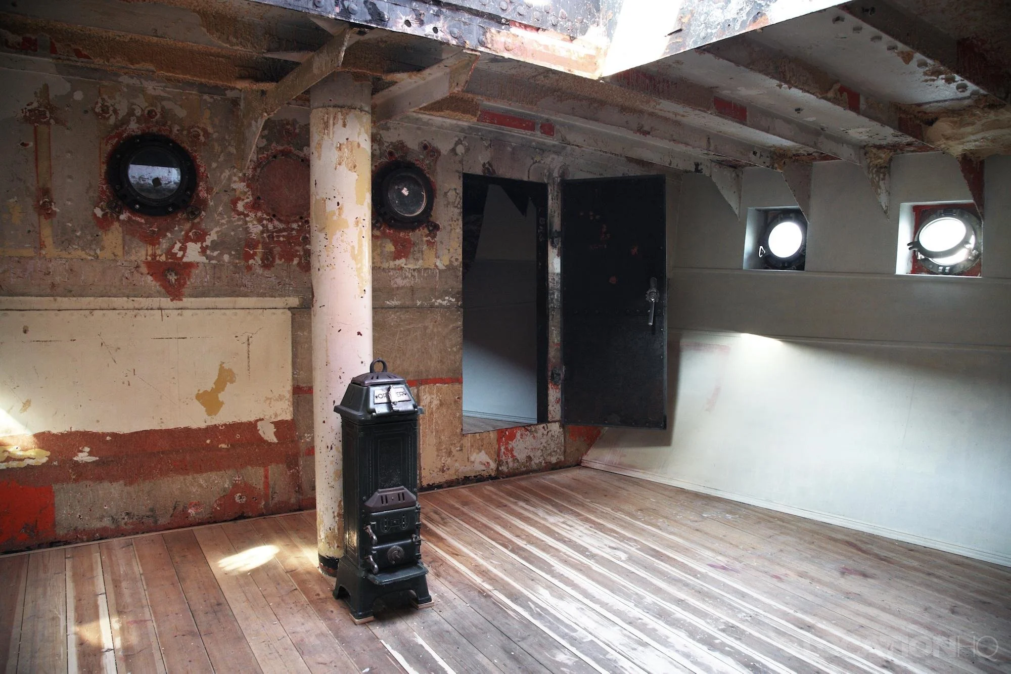 An interior of a rustic, unfinished room with wooden floors, exposed red brick walls and a white-painted wall on the right side. There are three small, round windows on the right wall, one of which is open. The ceiling is open with visible beams, and