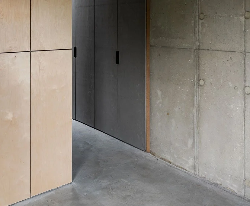 Photo of a modern interior corner with beige wood panels on the left, black sliding doors in the middle, and an unfinished concrete wall on the right, with concrete flooring.