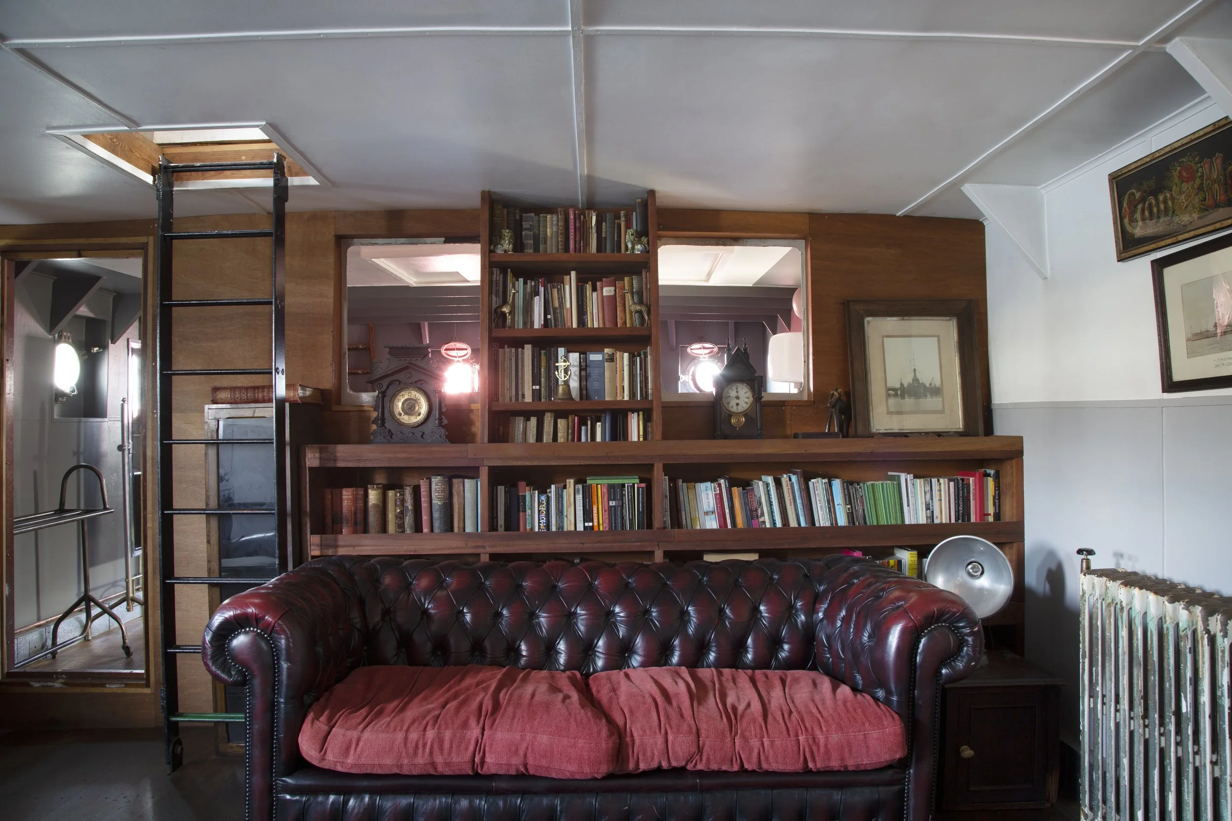 Interior view of a cozy living room with a leather Chesterfield sofa with a red cushion, a built-in wooden bookshelf filled with books, vintage clocks, and framed artwork, complemented by a white wall and a radiator.