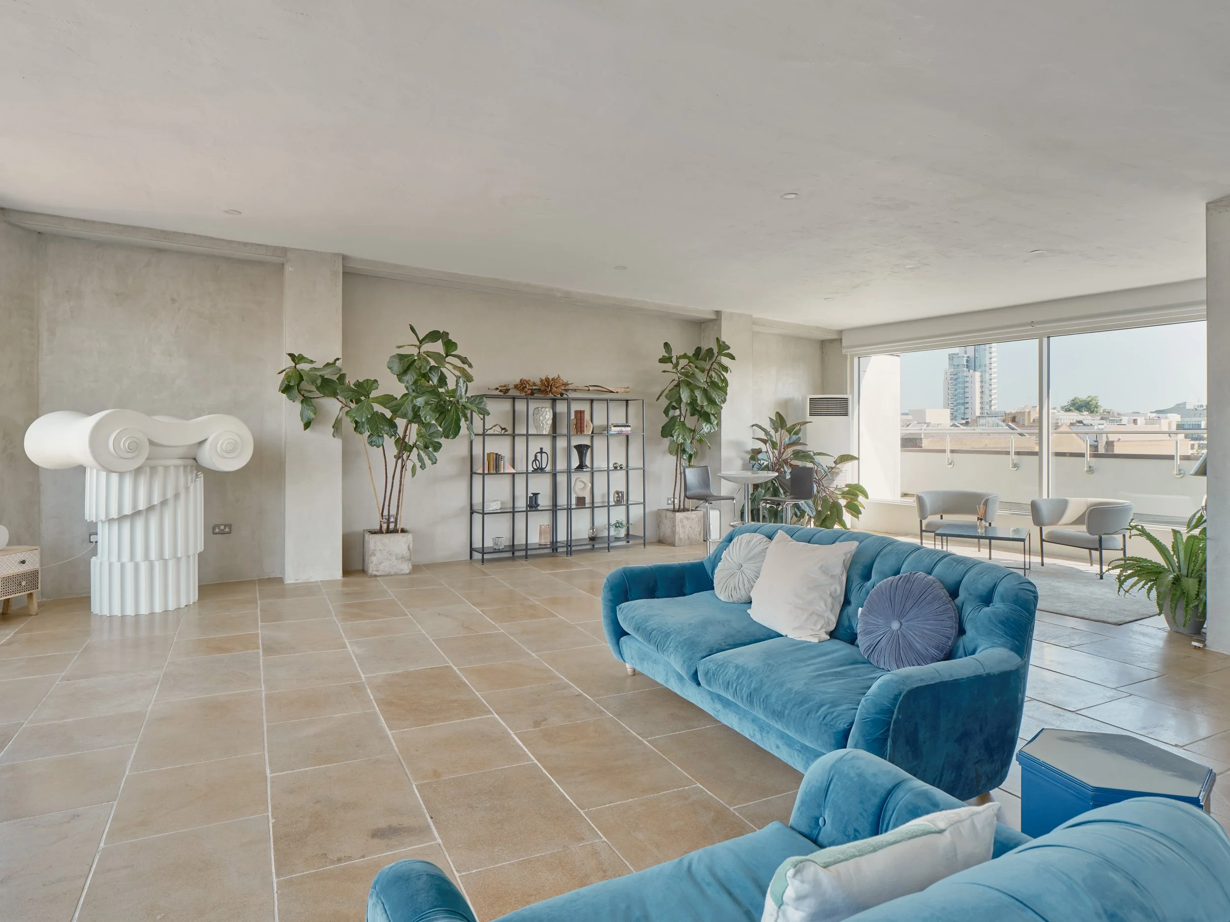 Modern living room with beige tiled floor, blue velvet sofas with white and purple pillows, a black metal shelving unit, large potted plants, a small round table, lounge chairs near the window, and cityscape view through large windows.