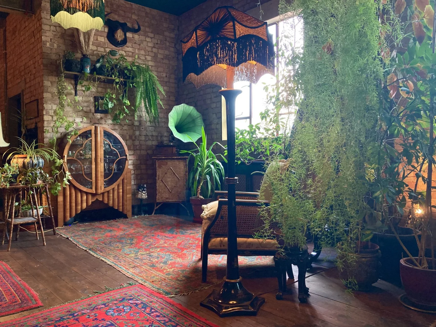 A cozy indoor space with vintage furniture, lush green plants, a brick wall, and a colorful patterned rug. Natural sunlight filters through large windows, illuminating the room.