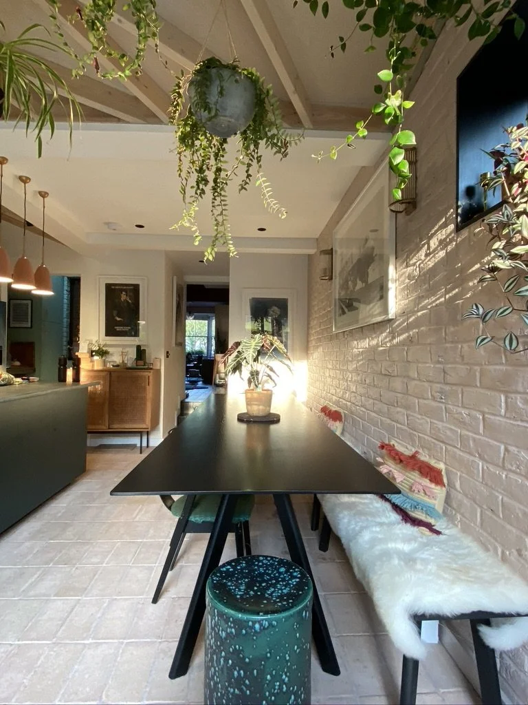 Interior of a modern dining area with a black table, colorful stools, a patterned bench with pillows, hanging plants, and a brick wall with a mounted TV.
