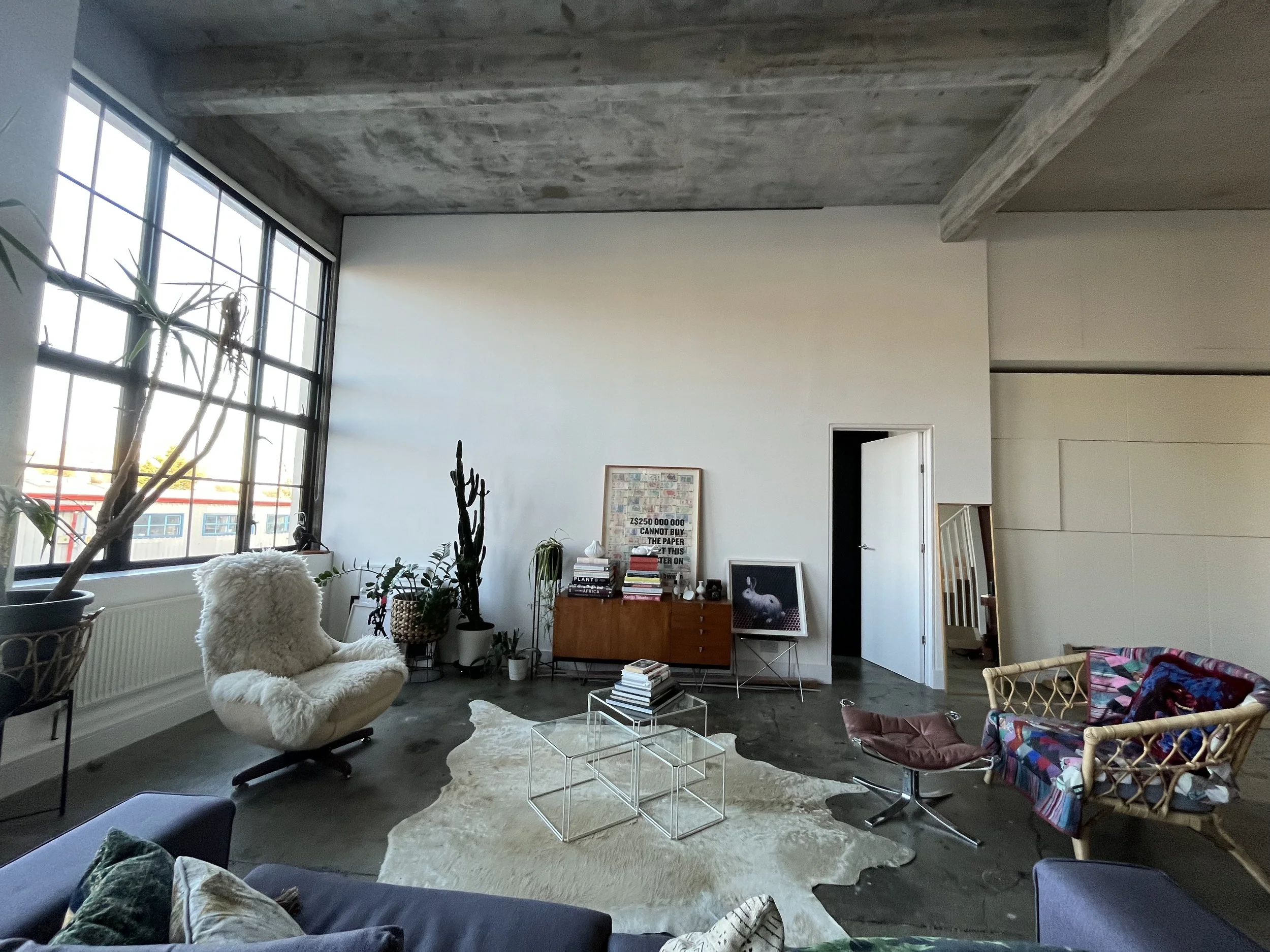 Modern living room with large window, potted plants, armchair with sheepskin, coffee table, patterned chair, and various decorative items.