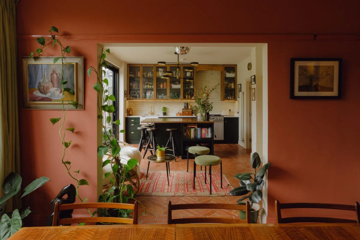View of an open-concept kitchen and dining area with a reddish-pink wall, wooden floor, and various plants. The kitchen has glass-front cabinets, a black breakfast bar with stools, and a bookshelf. The dining area has a table and chairs, with framed 