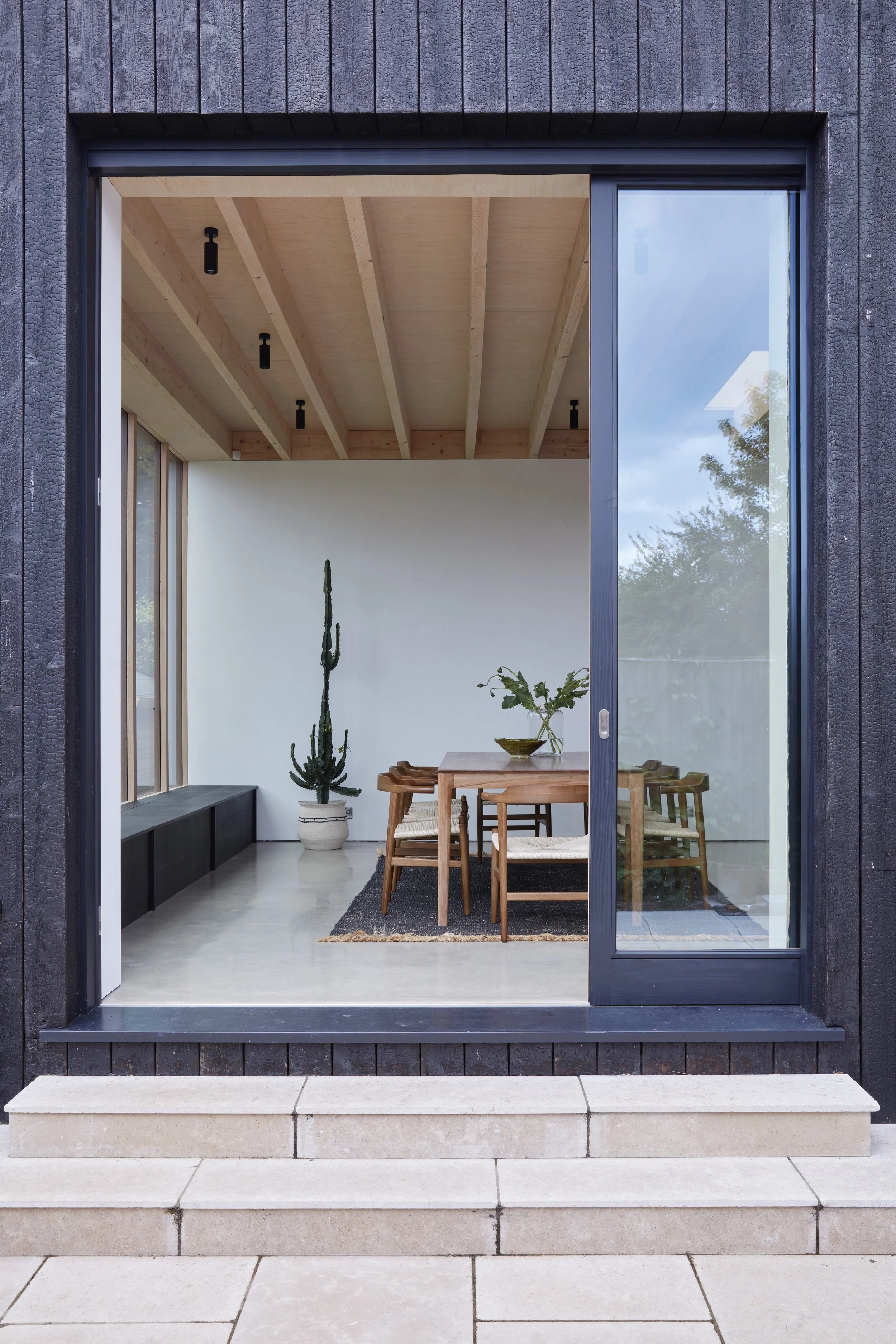 Interior view of a modern dining room with wooden table, chairs, potted cactus, and plant, seen through a large glass sliding door, with steps leading up to the entrance.