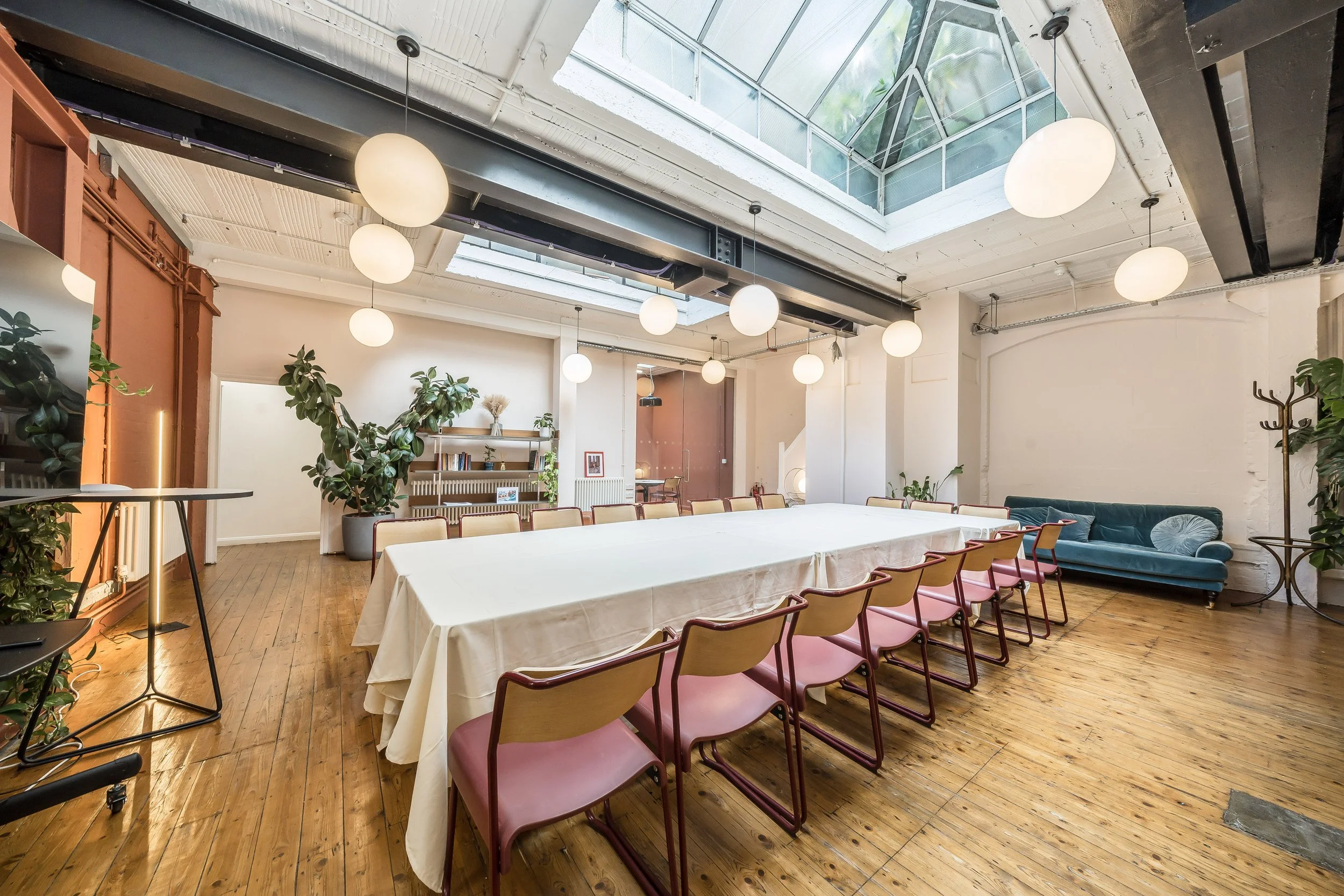 An airy conference room with a long table covered in a white tablecloth, surrounded by pink chairs, large indoor plants, a blue sofa, and decorative pendant lights hanging from a high ceiling with a skylight.
