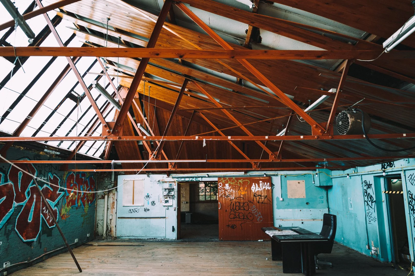 Interior of an abandoned building with graffiti on the walls, exposed wooden roof structure, and a single black chair near a small table.