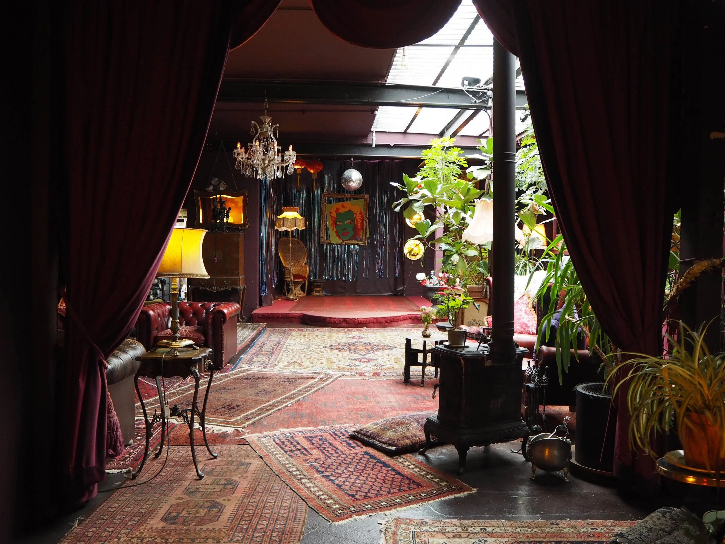 Eclectic vintage lounge with a stage, chandeliers, Persian rugs, vintage furniture, plants, and artwork, viewed from behind velvet curtains.