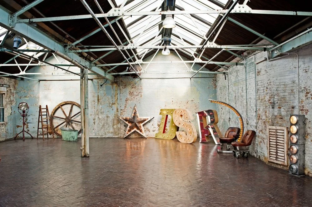 An industrial-style room with exposed brick walls, a high ceiling with metal beams and skylights. Large illuminated letters spelling 'LSR' and various vintage decor items including a star, a wheel, a ladder, a headlamp, and chairs.
