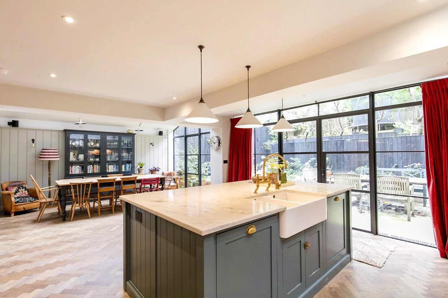Open-concept kitchen and dining area with large glass doors, navy blue kitchen island with white marble countertop, and a wooden dining table with chairs. Red curtains, a black glass cabinet, and outdoor patio furniture visible through the windows.