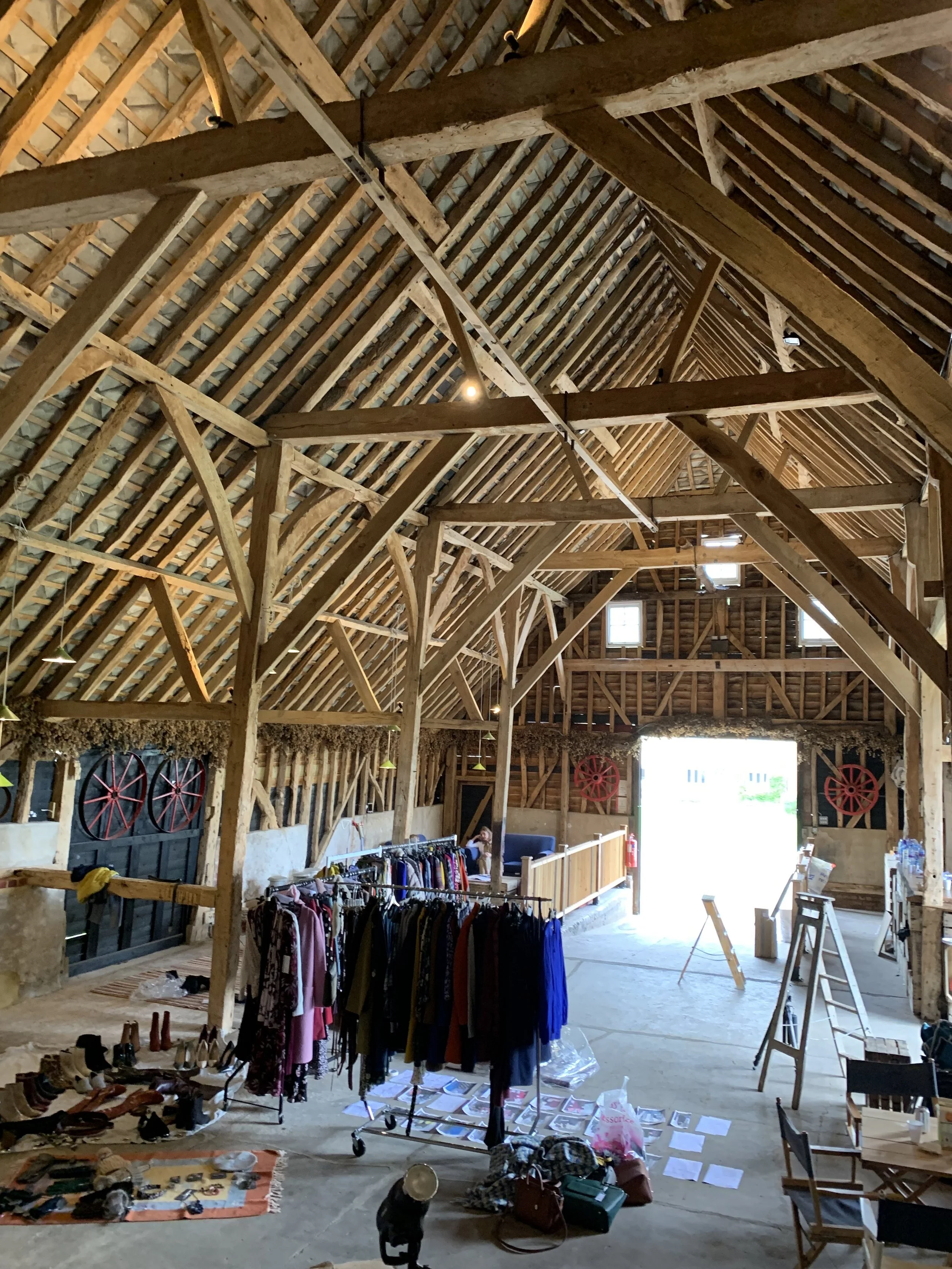 Interior of a rustic barn used as a thrift store with clothing racks, shoes, and various items for sale, with visible wooden beams and high ceilings.