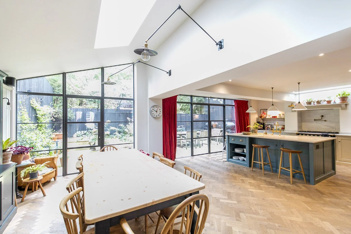 Bright kitchen and dining area with large windows, wooden dining table, plants, and red curtains.