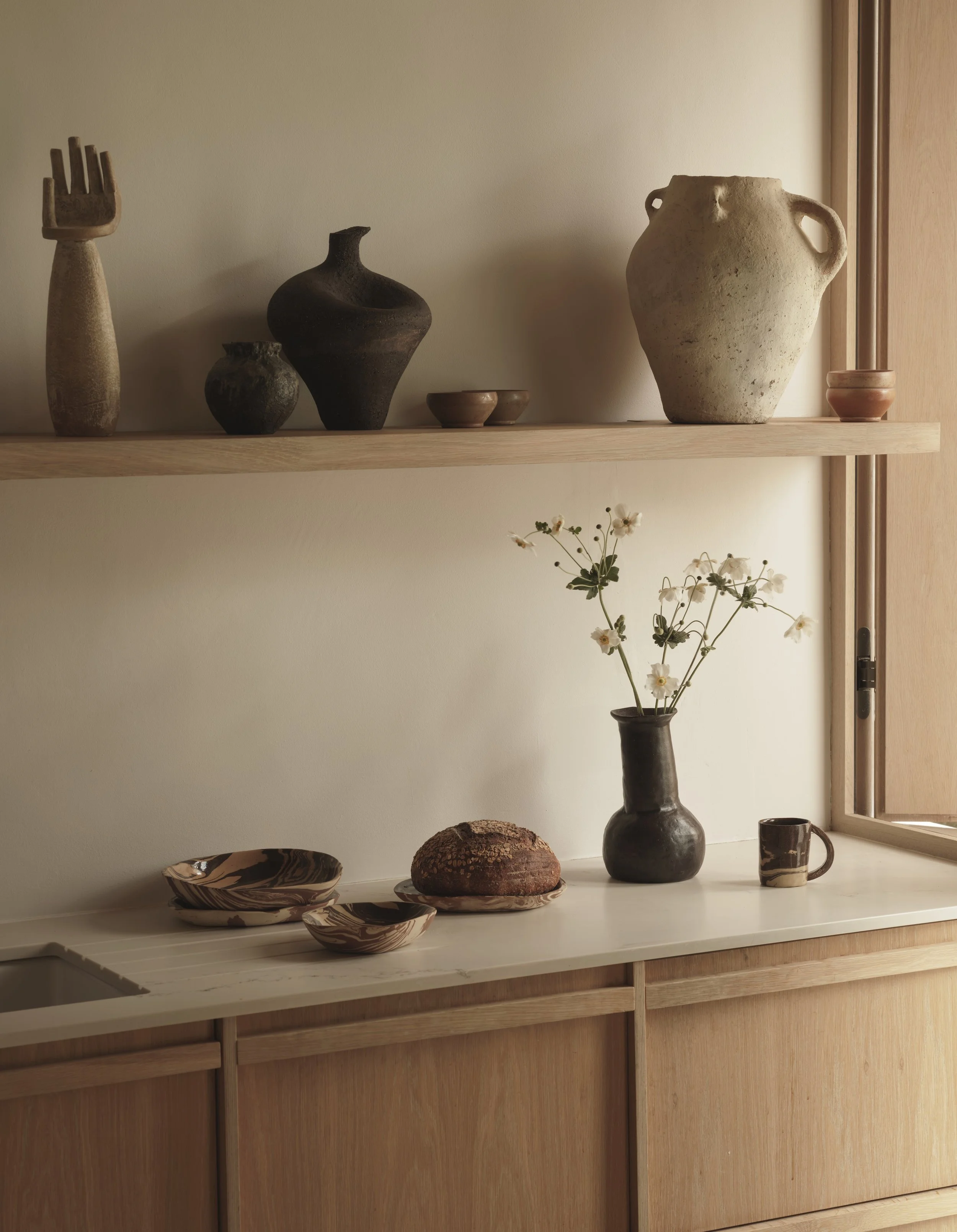 Interior shelf with ceramic vases, bowls, flower vase with white flowers, and a loaf of bread, against a light wall and wooden furniture.