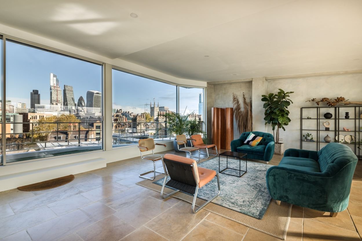 Modern living room with large windows overlooking a city skyline, featuring teal and tan furniture, a metal bookshelf, and green plants.