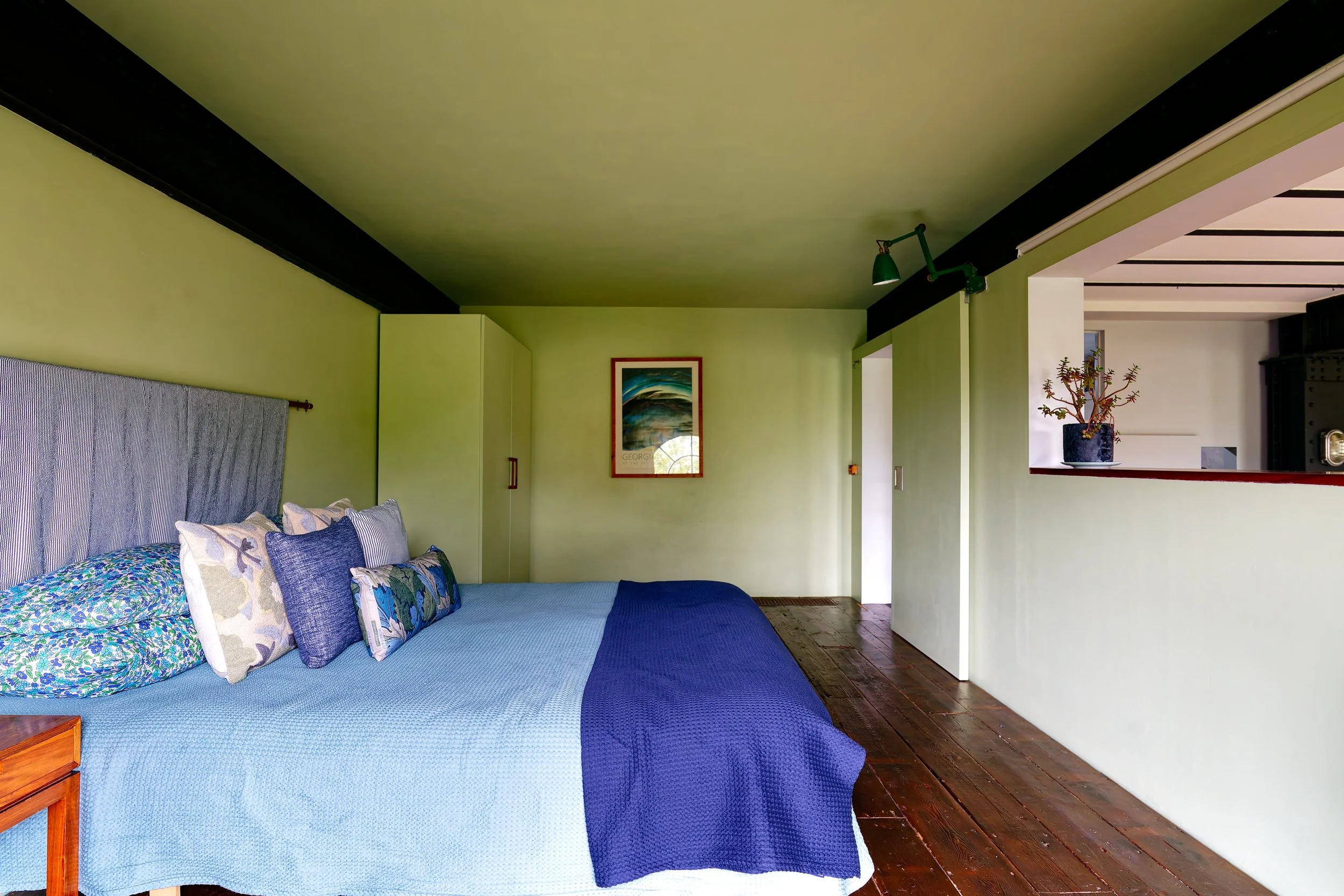 A bedroom with a green wall and dark wooden floor, a bed with blue linens and multiple pillows, a small wooden nightstand, and a framed picture on the wall. There is a partial view of a kitchen with a potted plant on a ledge.