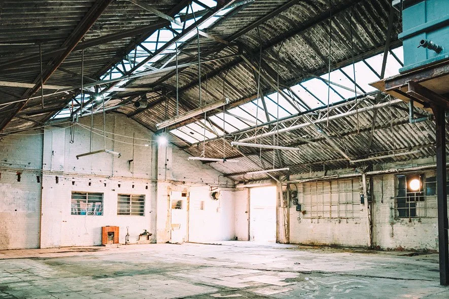 Empty industrial warehouse with high ceiling, metal roof, and exposed structural elements, illuminated by natural light through skylights.