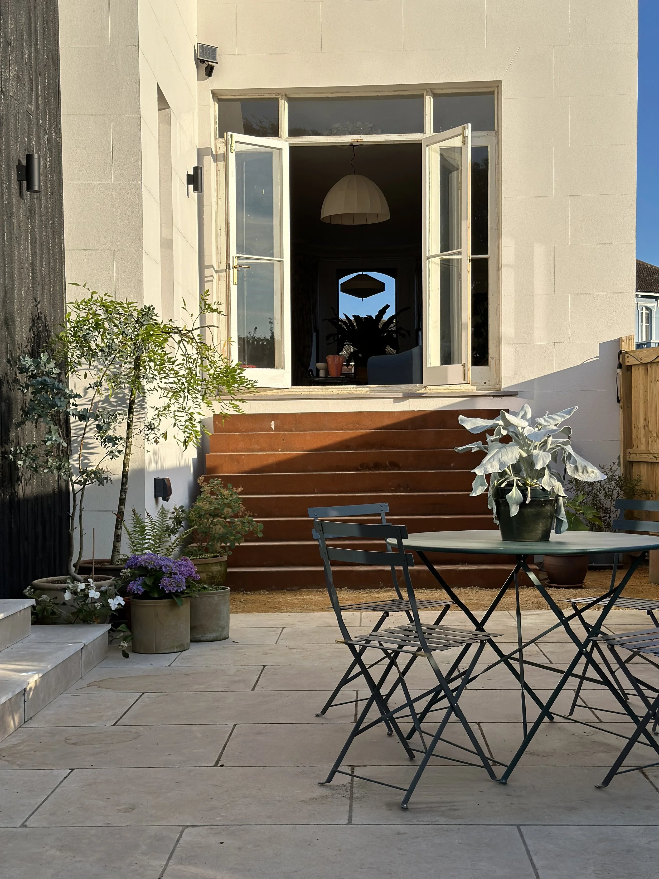 An outdoor patio area with a round table and four black chairs, potted plants, and steps leading up to a house with open white French doors, inside a living room with a hanging light and houseplants.