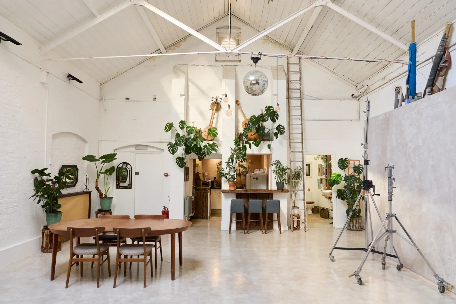 Interior of a bright, spacious room with white walls and ceiling, featuring a wooden dining table with six chairs, various potted plants including monstera, guitar decor on the wall, a disco ball hanging from the ceiling, and photography equipment se