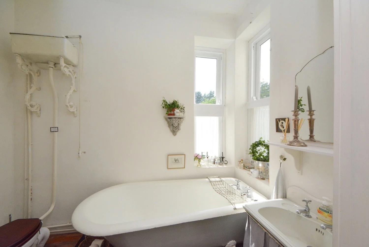 Bright vintage bathroom with antique white bathtub, window with white curtains, and decorative shelves with plants and candlesticks.