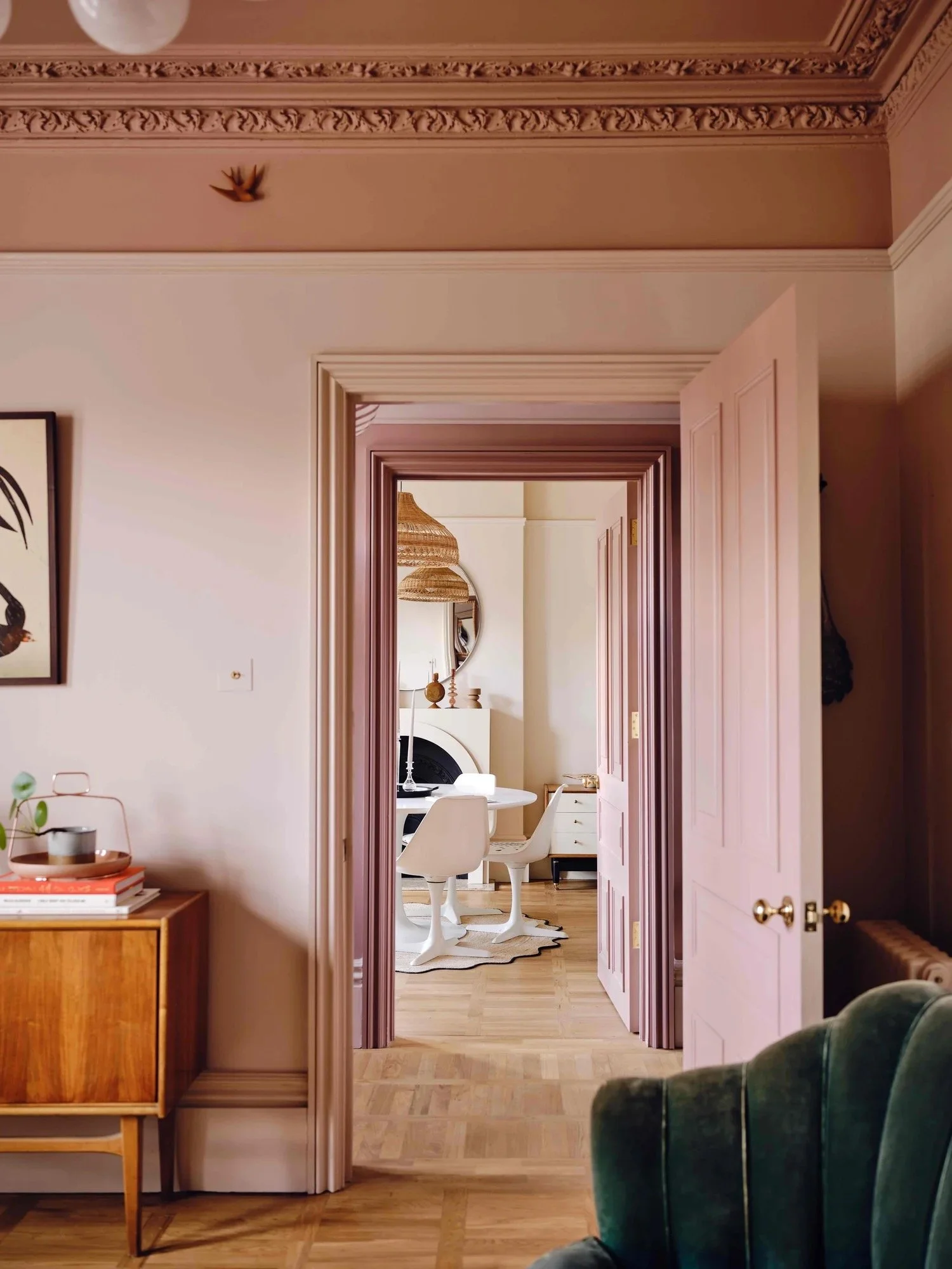 View of a room with pink doors open to a dining area with white chairs, a round mirror, and a fireplace in the background.