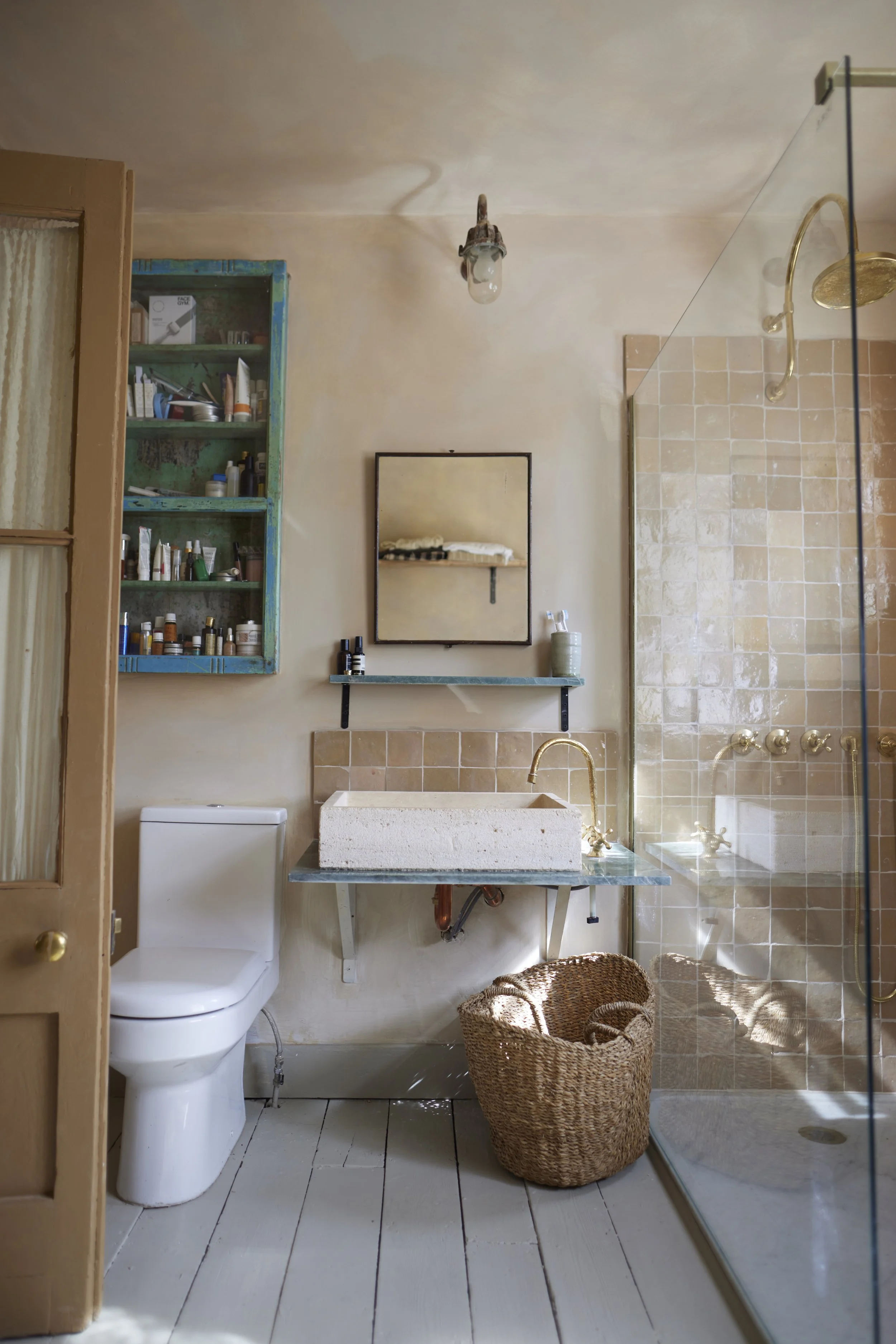 A rustic bathroom featuring a white toilet, a concrete sink with a gold faucet, a small mirror, a green shelf with toiletries, a wicker laundry basket, and a glass-enclosed shower with beige tiled walls.