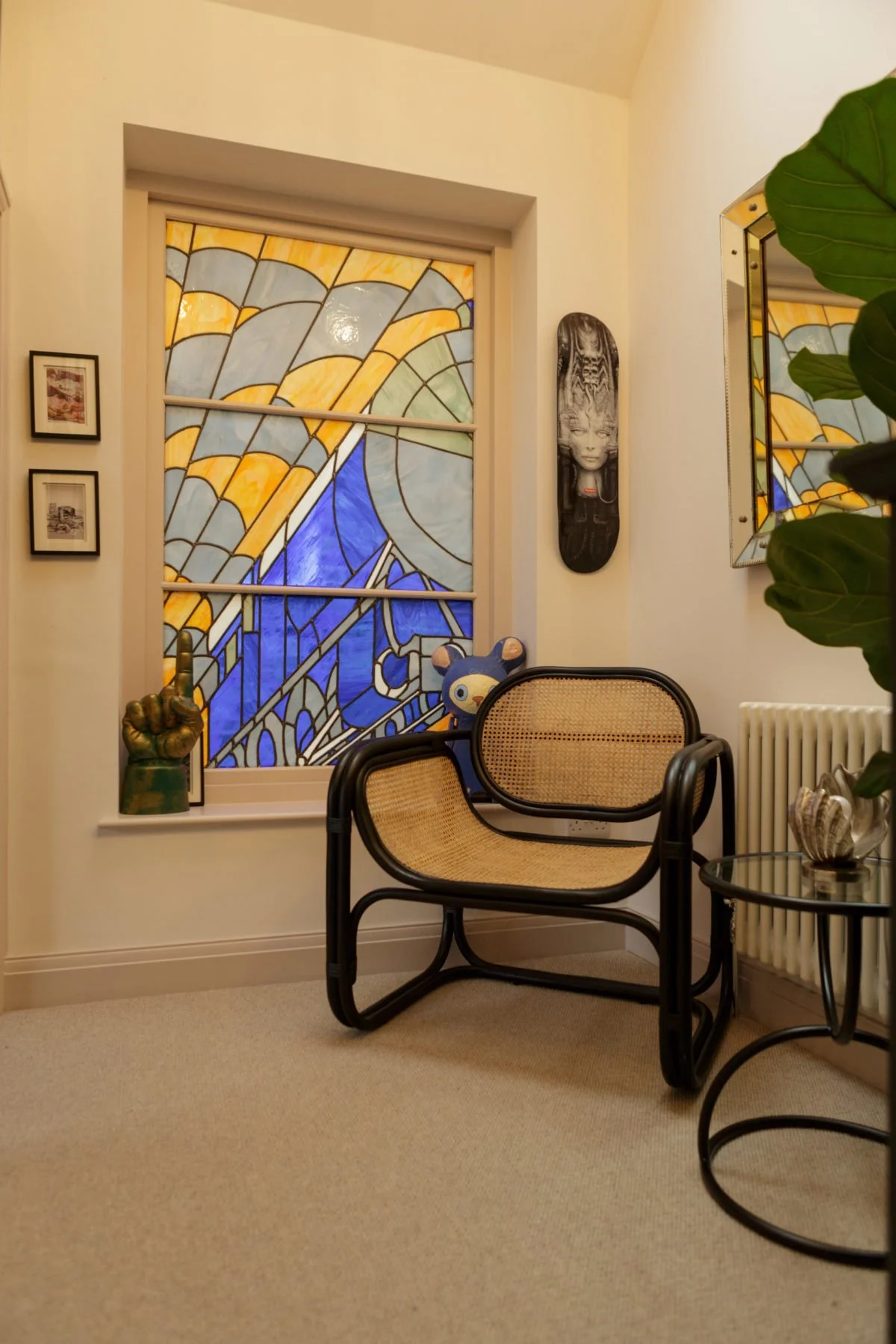 A cozy room corner featuring a stained glass window with abstract yellow, blue, and green shapes, a wicker-back armchair with a black frame, a round side table with seashell decorations, tropical plant leaves partially visible, framed photos on the w