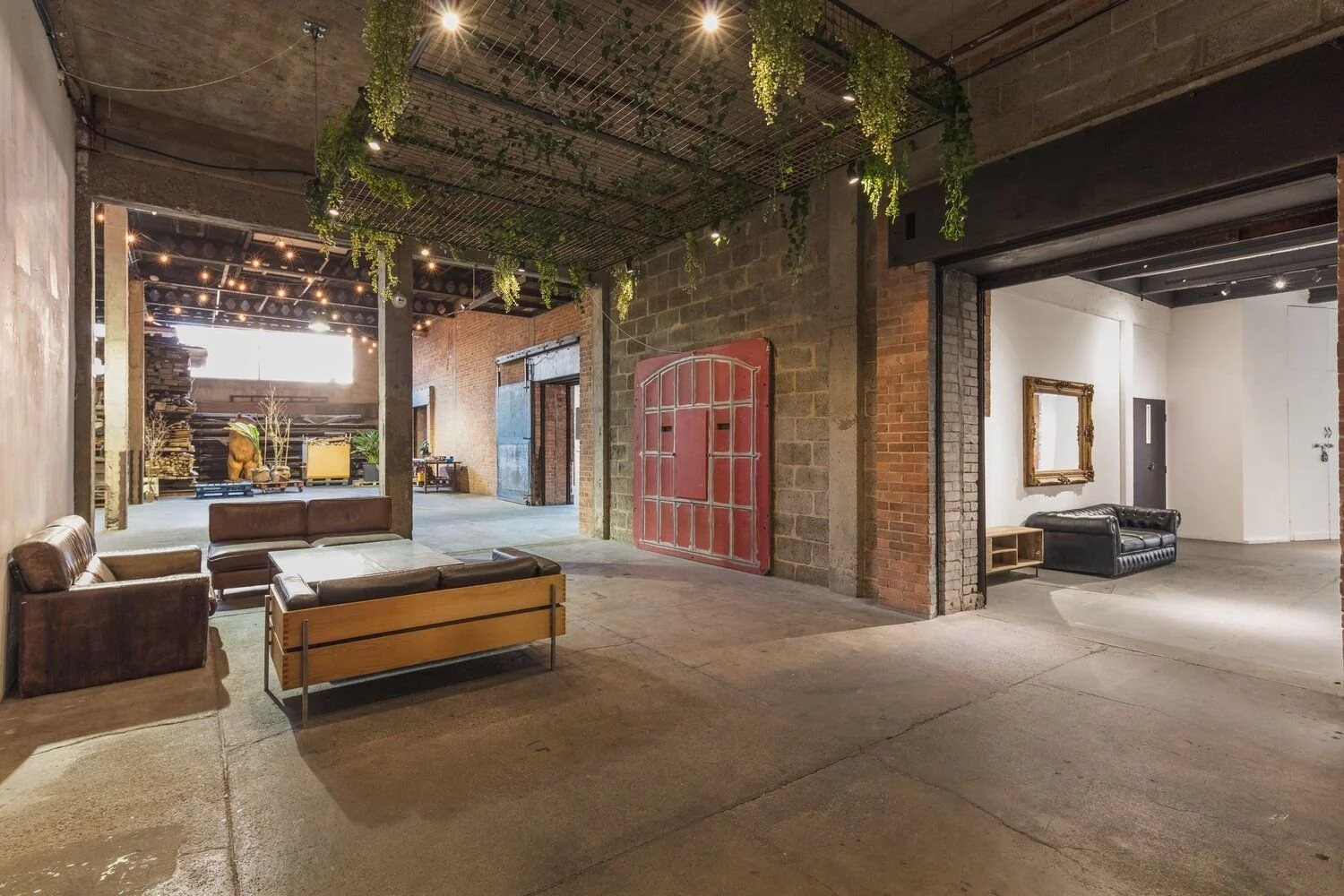 Interior of a modern industrial-style space with exposed brick walls, hanging plants, seating areas with leather sofas, and string lights.