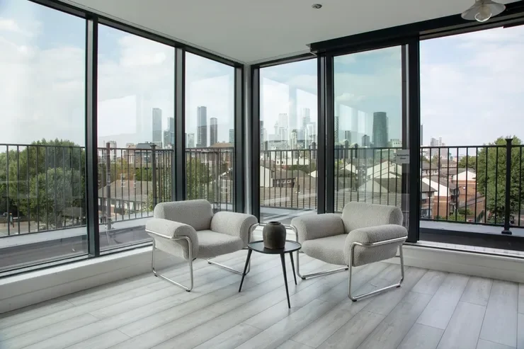Interior of a modern room with large glass windows showcasing a city skyline, two gray armchairs, a small black side table with a black vase, and a ceiling fan.