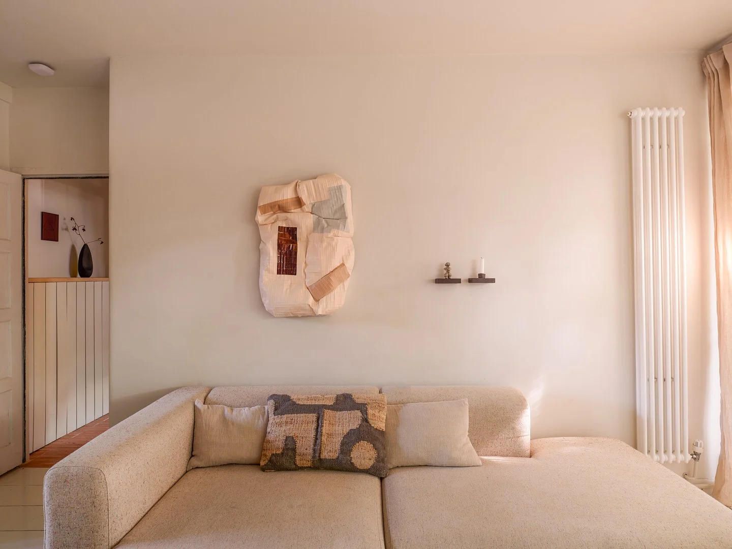 Living room with beige sectional sofa, beige pillow, abstract wall art, two small wall shelves with candle and figurine, radiator, curtains, and visible hallway with decorative vases.