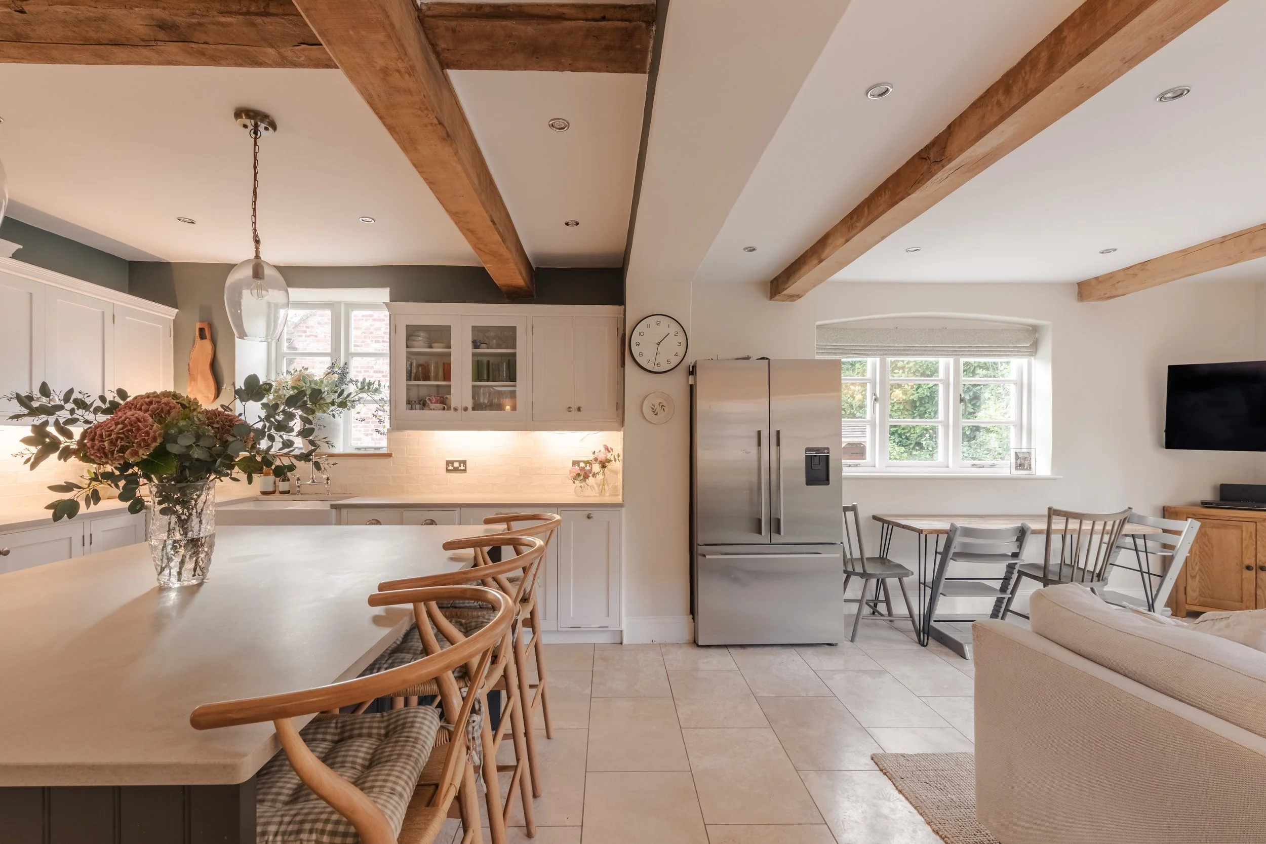 Open-concept living space with a kitchen and dining area, featuring white cabinets, a stainless steel refrigerator, a wooden beam ceiling, and large windows.