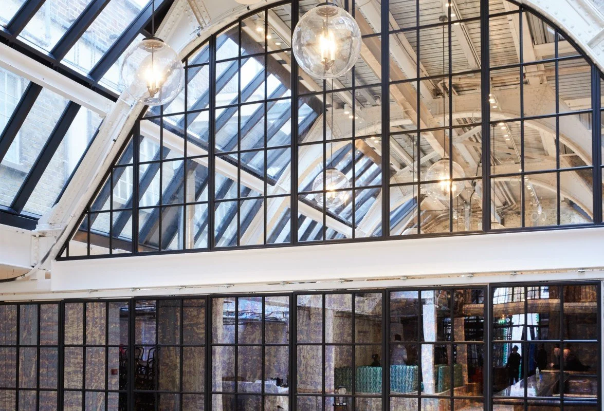 Interior of a modern industrial-style building with large glass windows and ceiling, multiple hanging light fixtures, and visible structural beams.